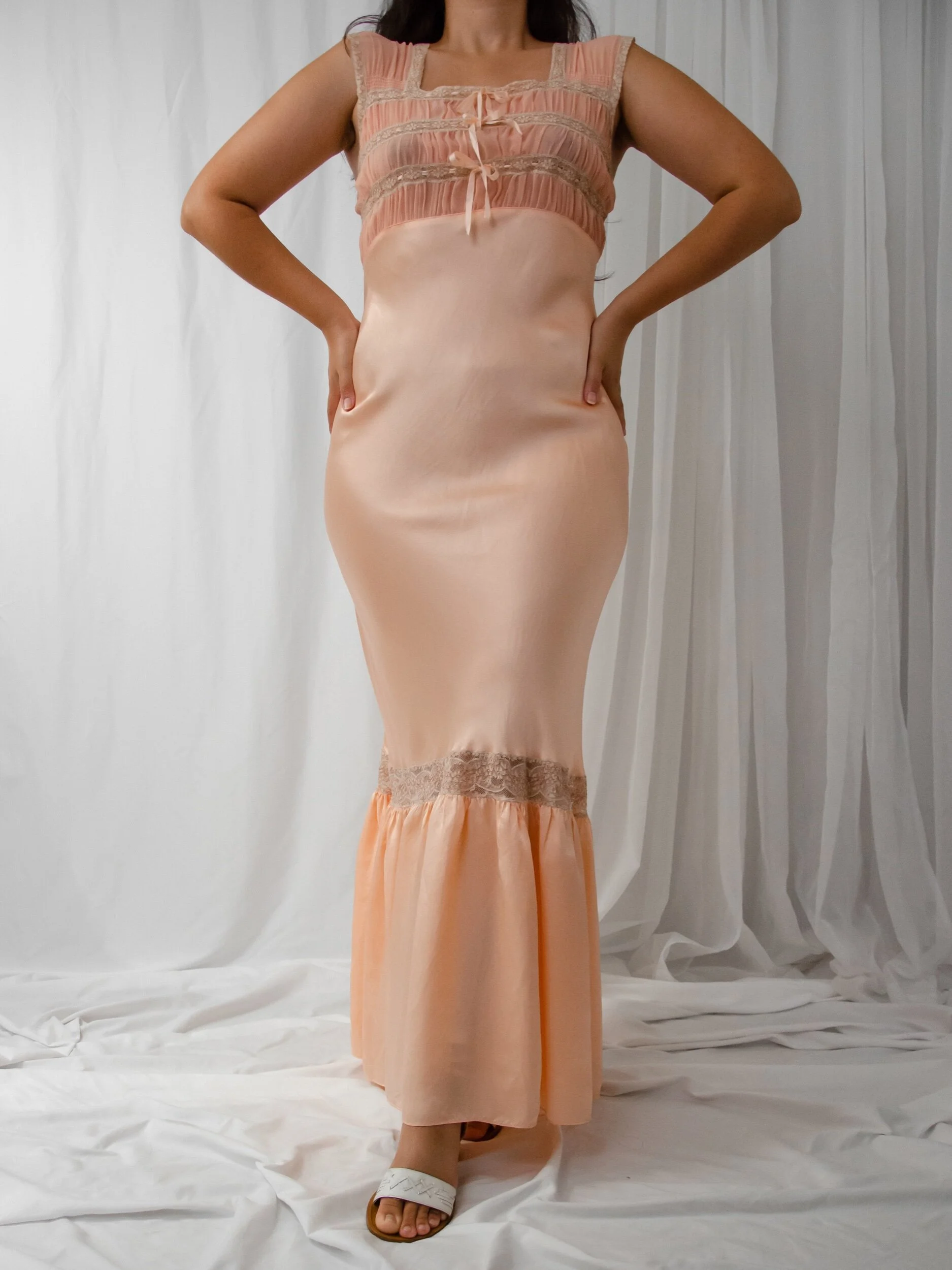 Front view of the 1930s peachy pink satin slip dress on a model with hands on hips.