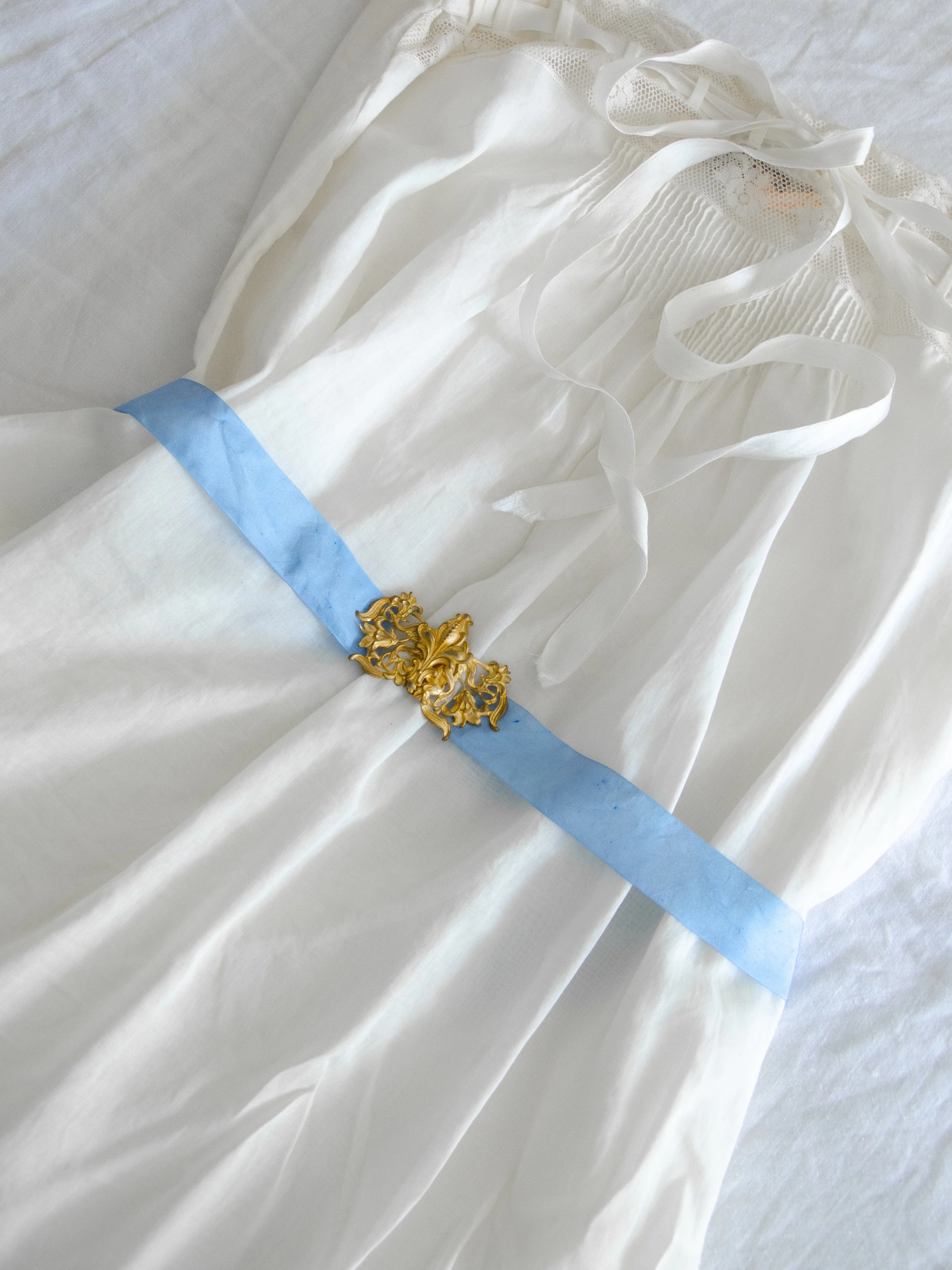 1930s blue silk ribbon belt with gold art nouveau buckle cinched around a white cotton slip, angled view.