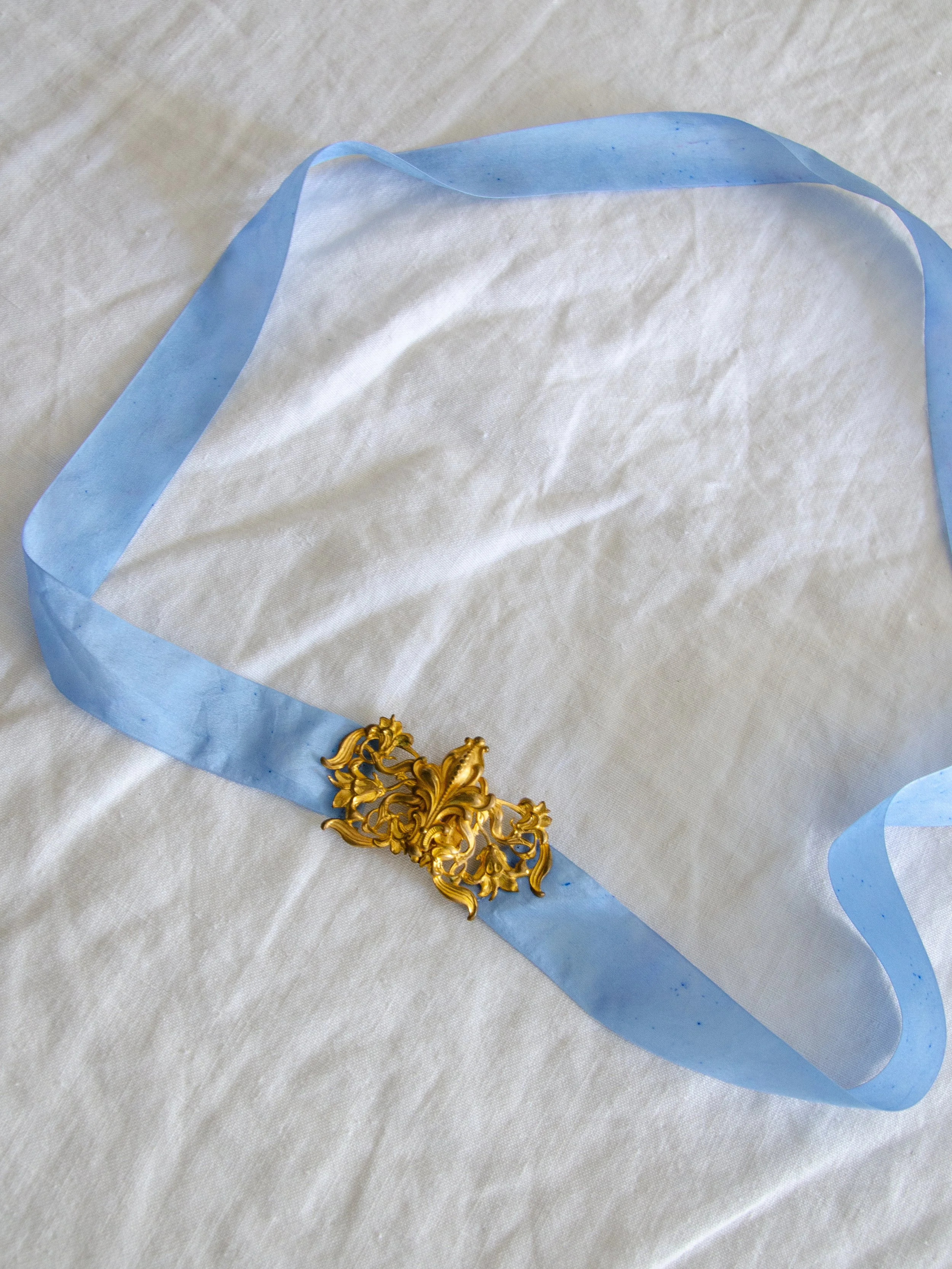 1930s sky blue silk ribbon belt with ornate gold buckle laid flat in a loop on white fabric.