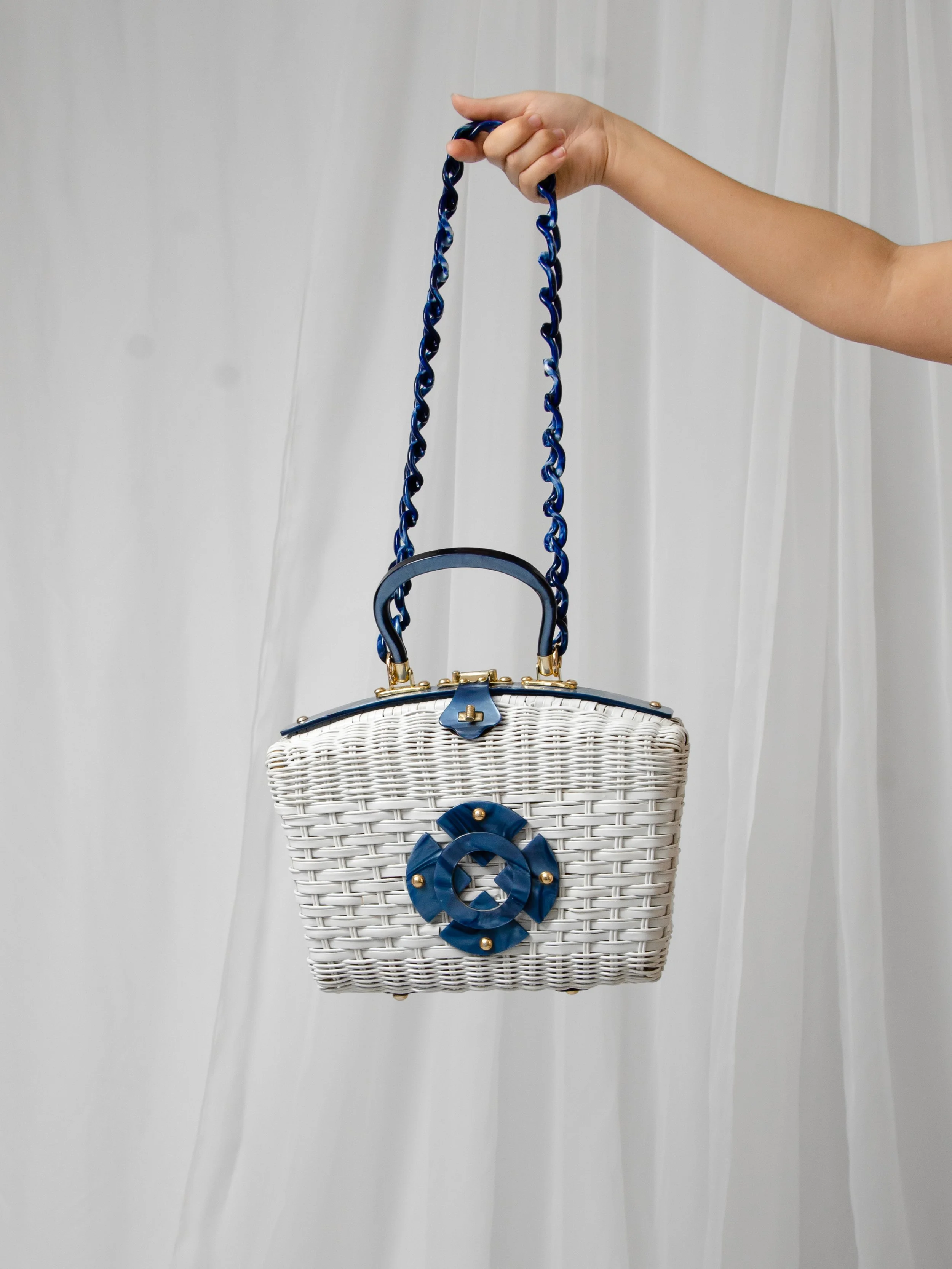 1950s white wicker purse with blue lucite top handle hanging from a marbled blue chain strap held by hand.