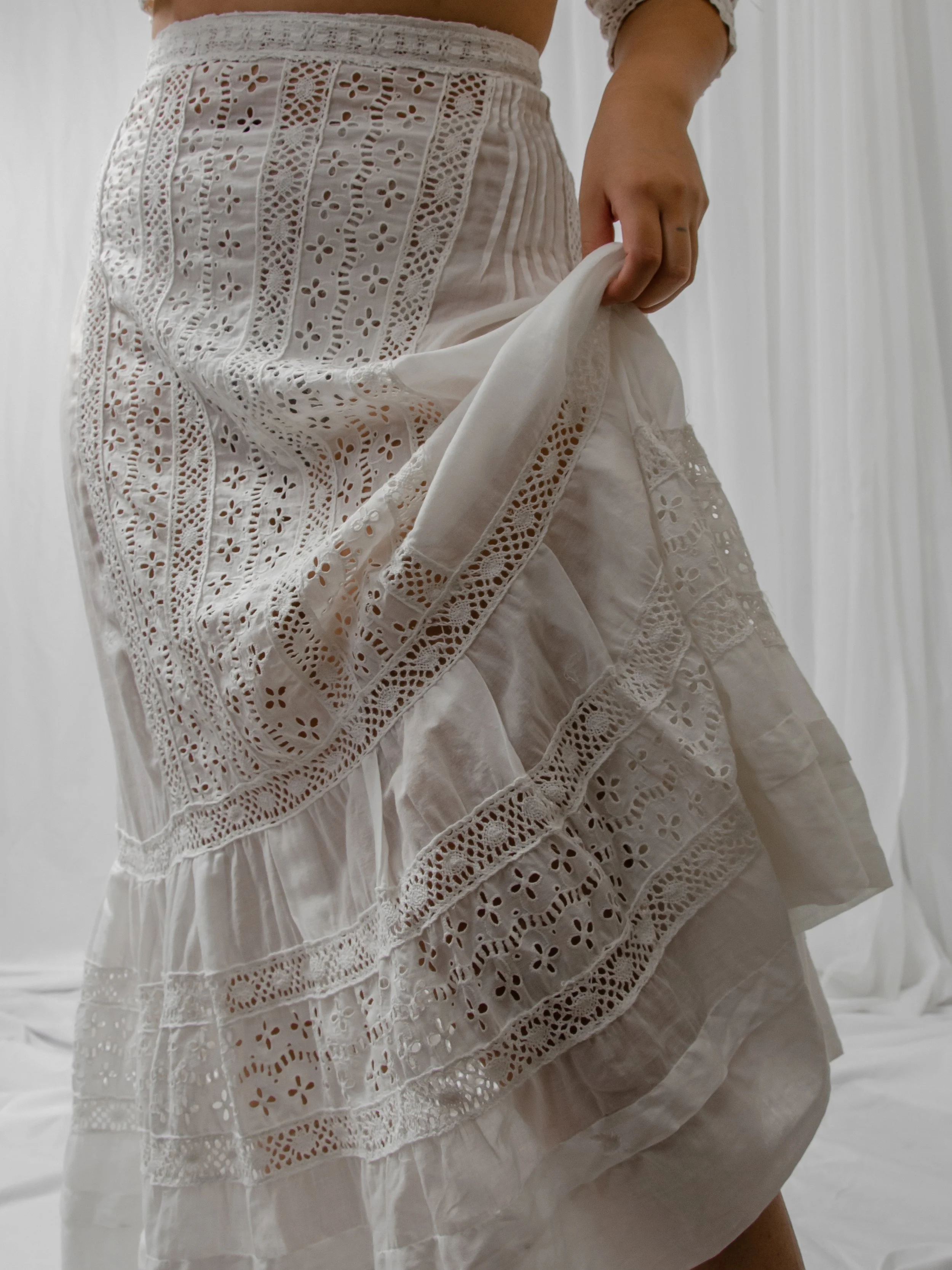 Detail of the Edwardian white eyelet skirt lifted by hand, showing tiered lace and eyelet panels.