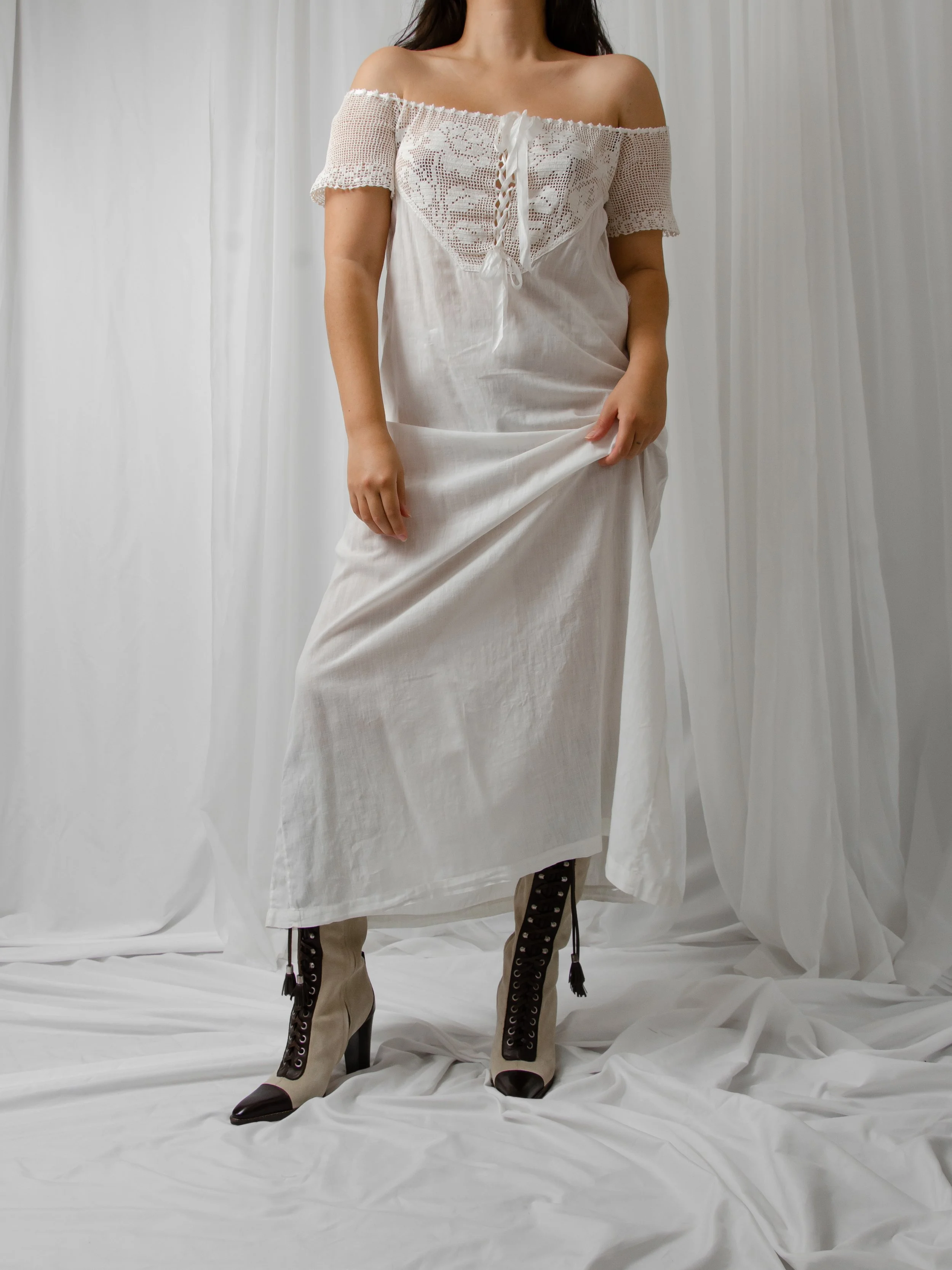 Edwardian white cotton off-shoulder dress on a model holding the skirt, showing lace-up boots.