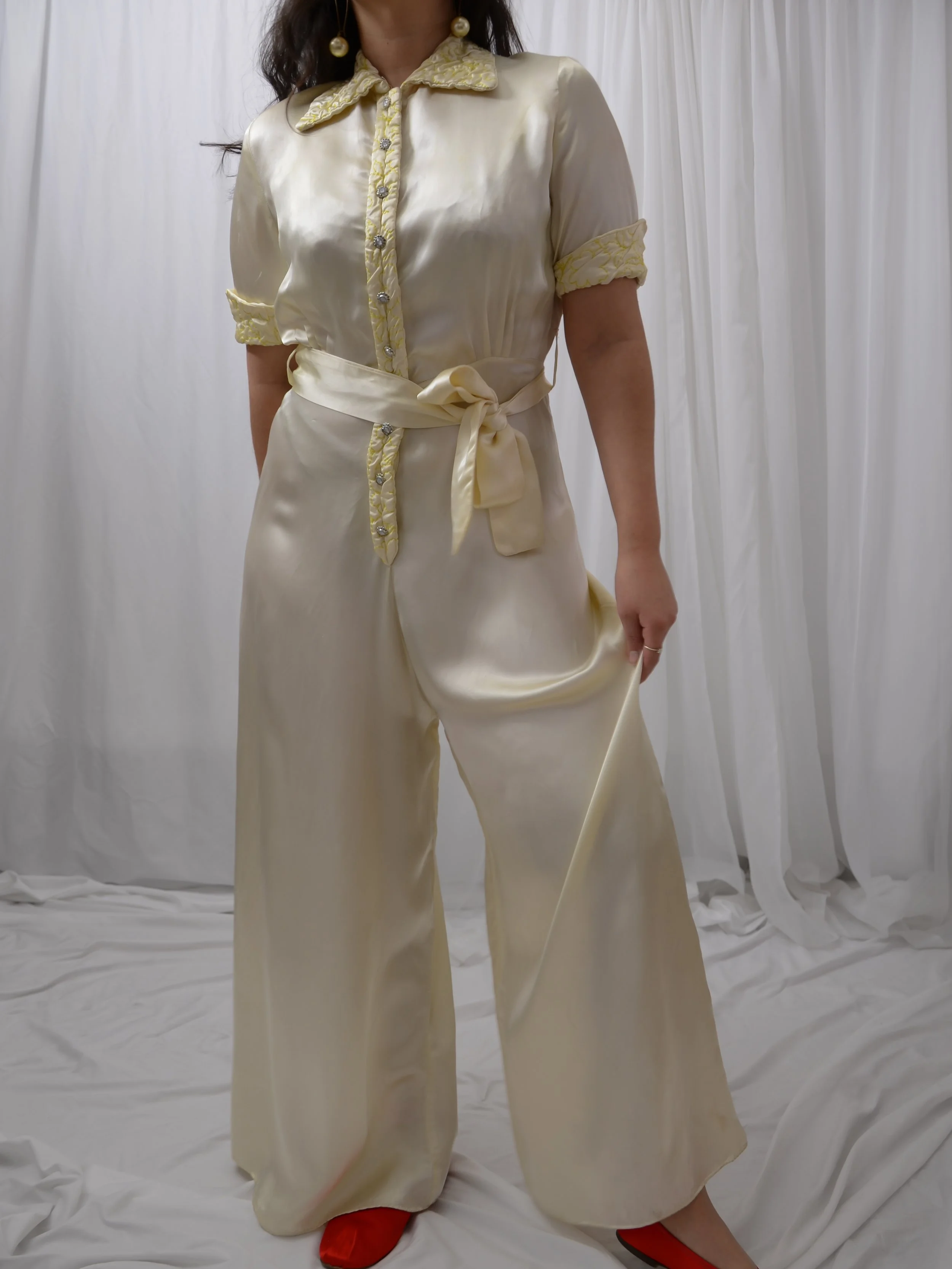 1940s buttercream satin jumpsuit on a model holding out one wide pant leg, with a tied sash.