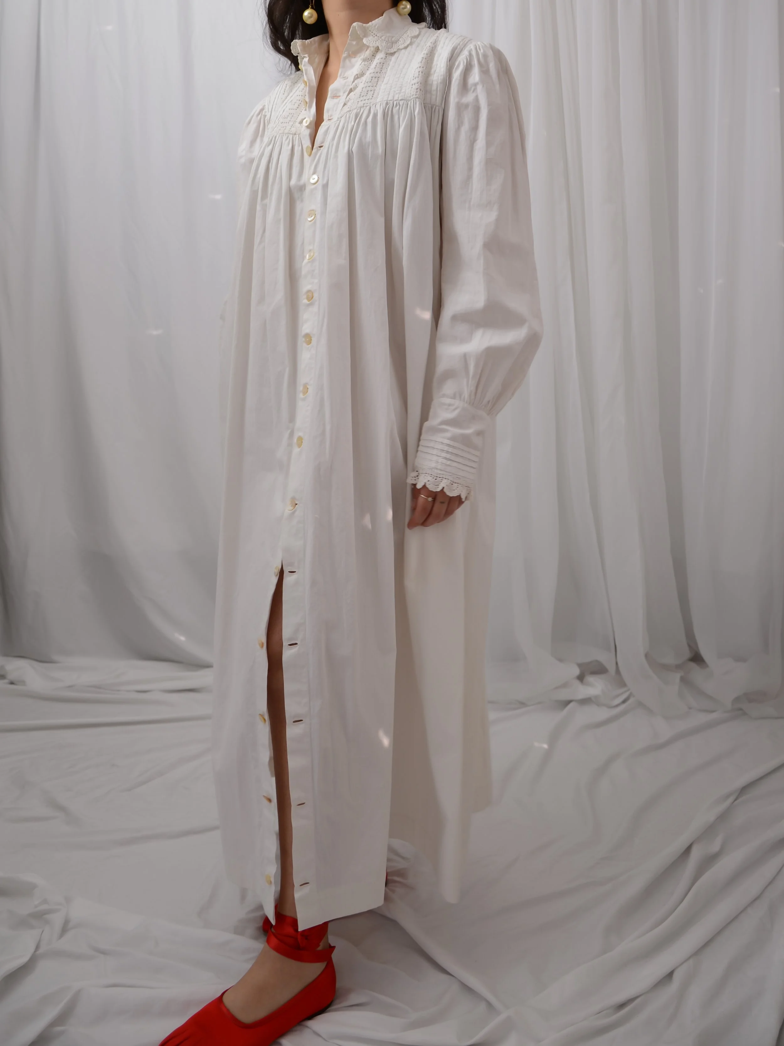 Antique white cotton button-down dress on a model, partly unbuttoned at the hem over red flats.
