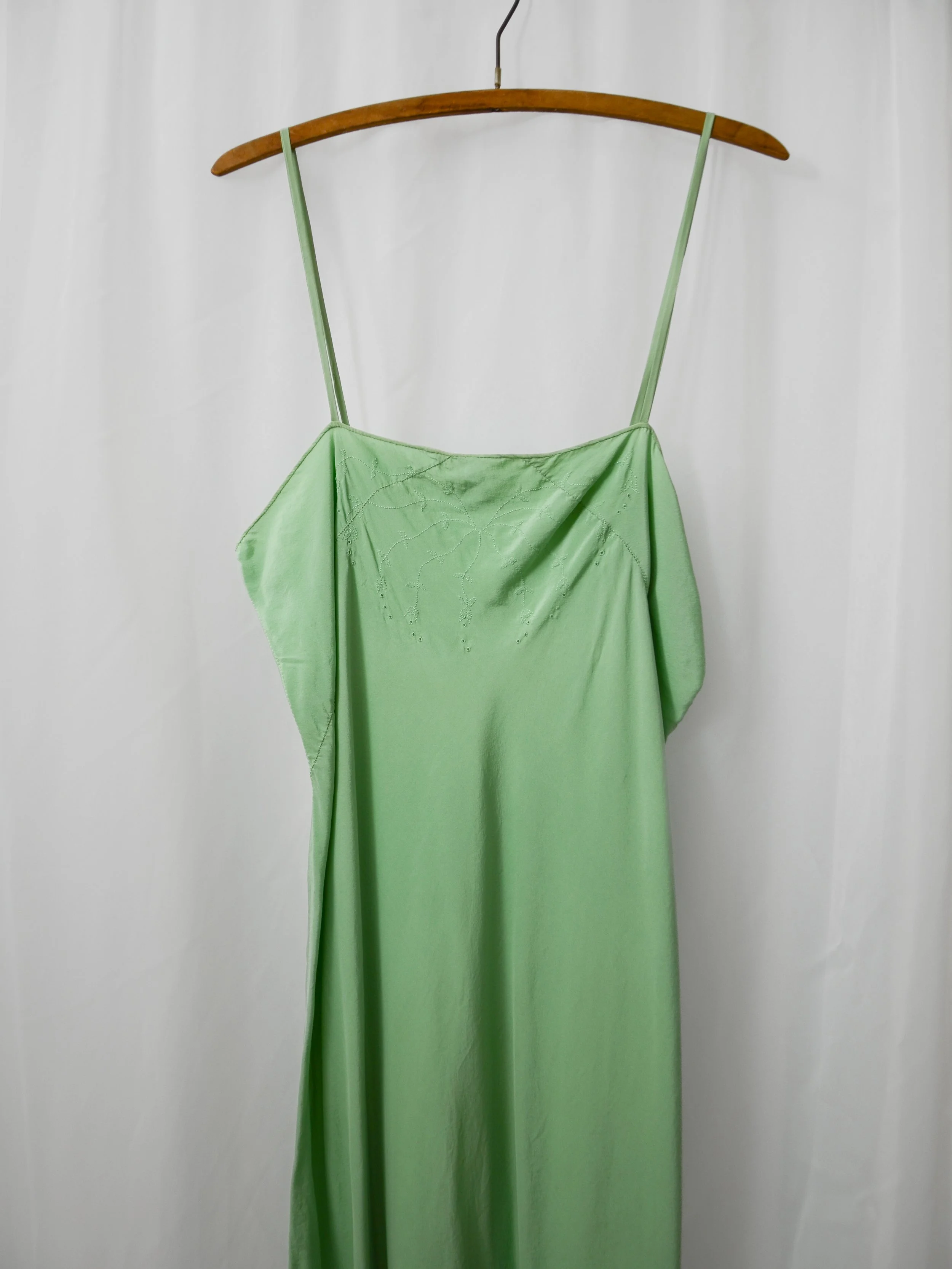 Close-up of the 1920s sage green silk slip dress on a hanger, showing vine and leaf stitching at the bust.