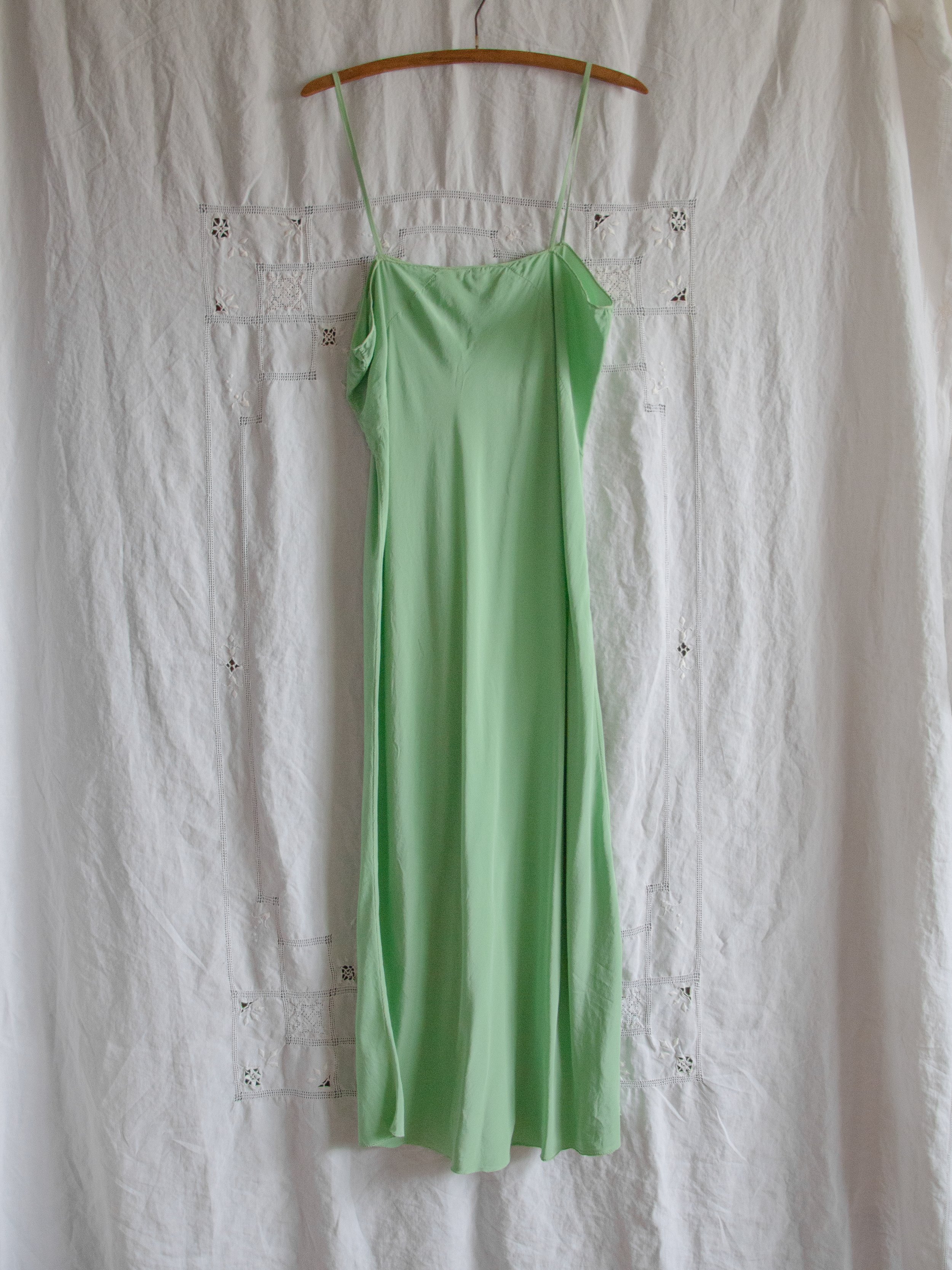 Late 1920s sage green silk slip dress on a wooden hanger against white linen, full-length view.