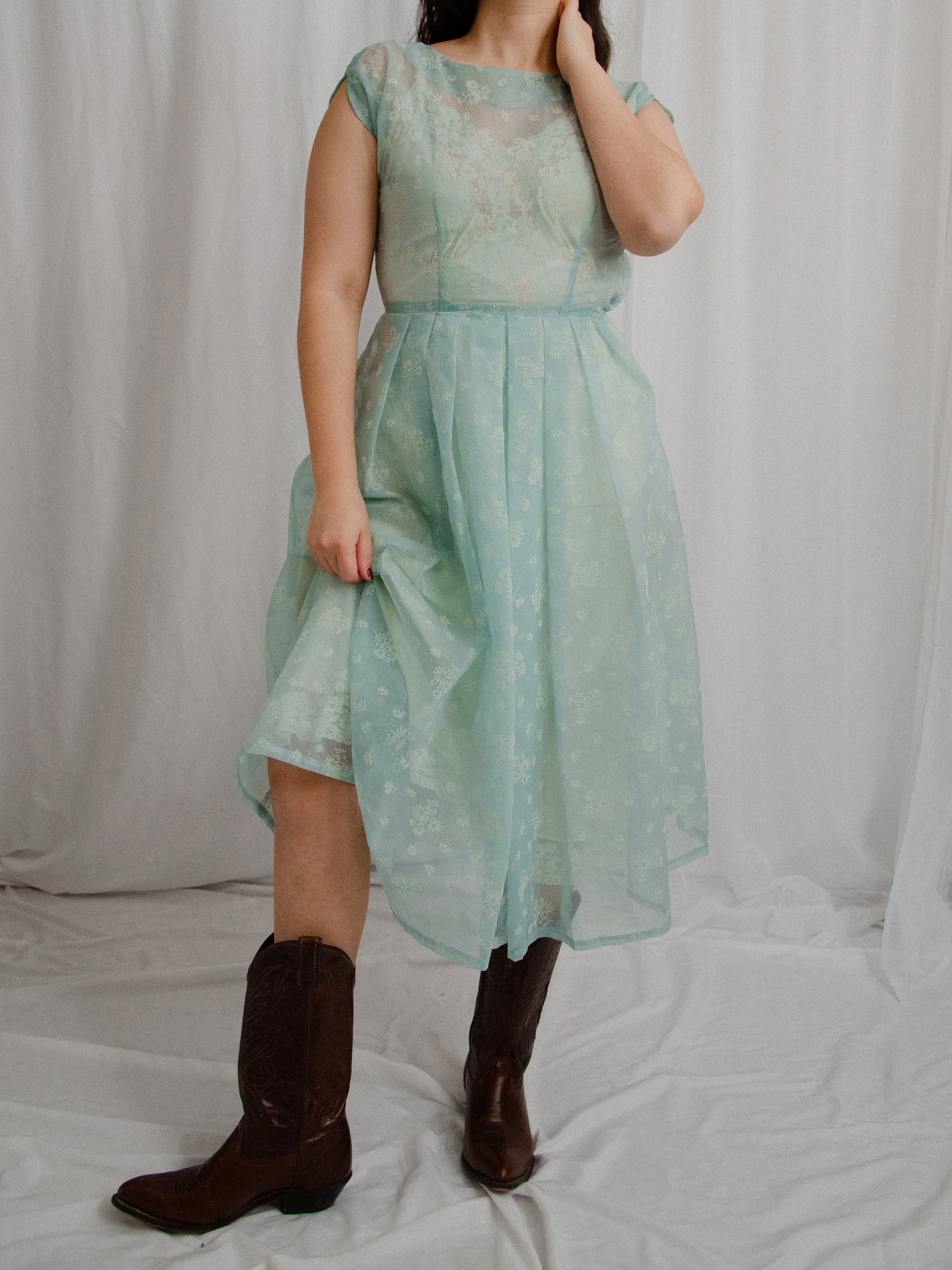 1950s sheer blue floral flocked dress on a model lifting the skirt to show its full pleated shape.