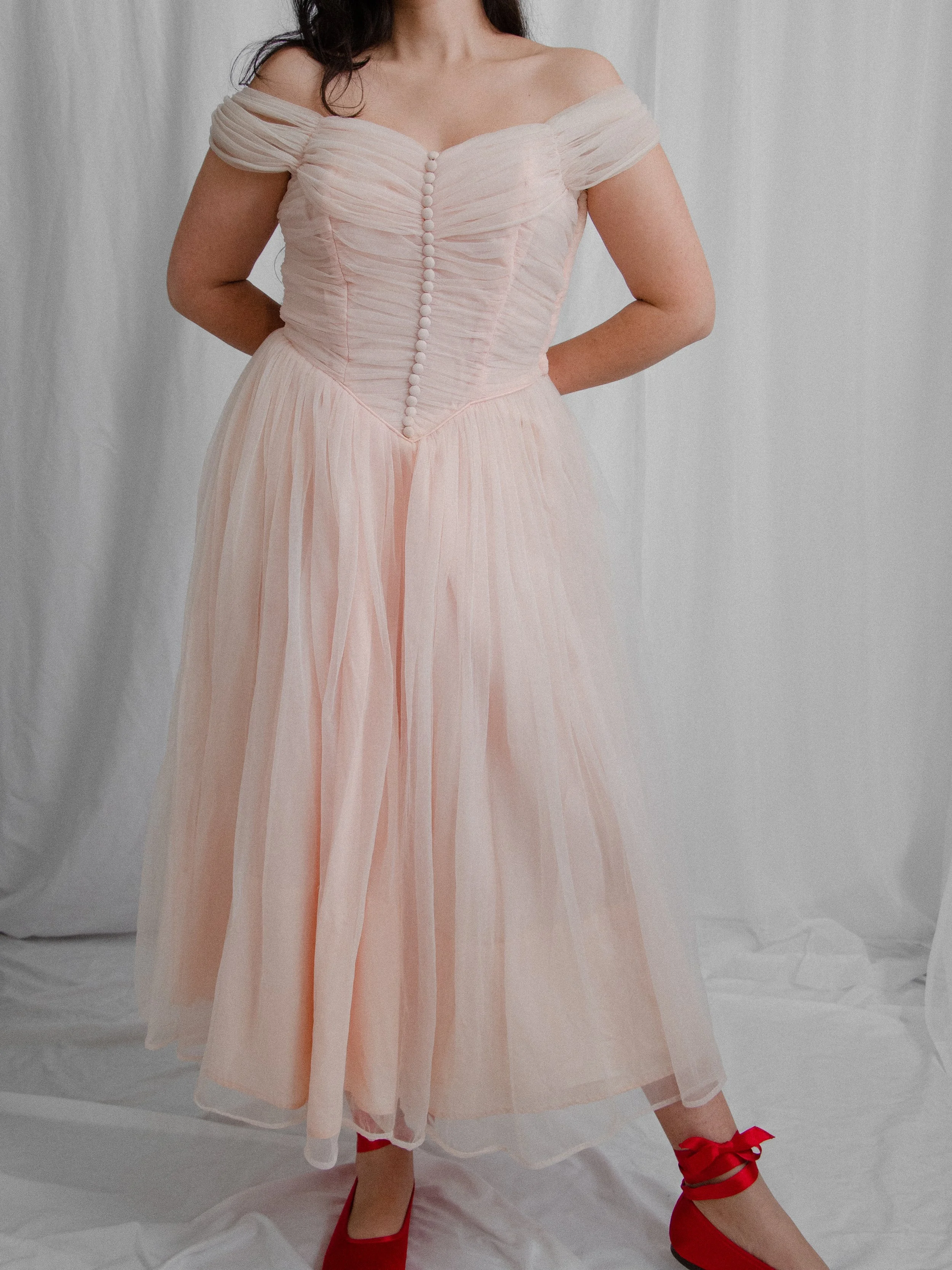 Front view of the 1950s pink ballerina dress worn off the shoulders on a model with red flats.