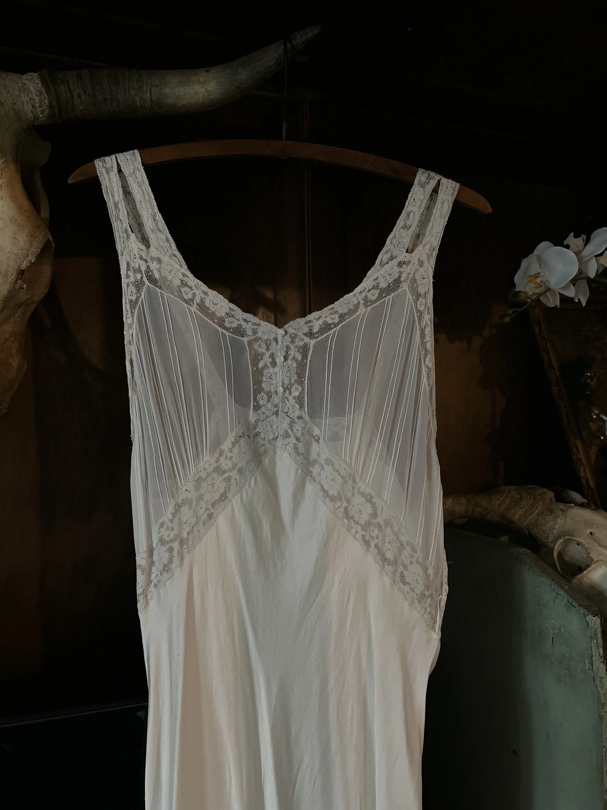 Close-up of the 1930s ivory slip dress bodice on a hanger against a dark background, showing pintucks and lace.