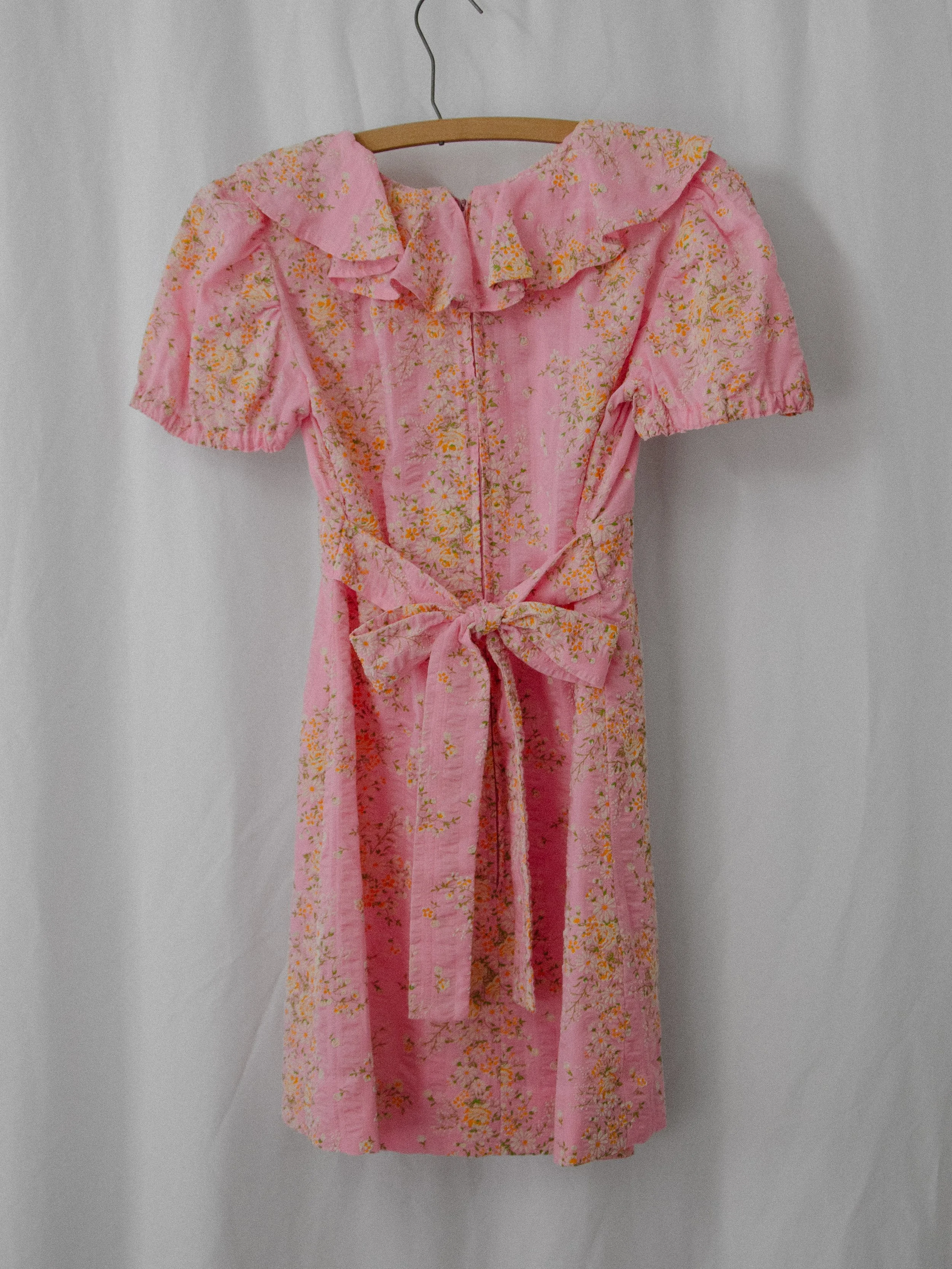 Back view of the 1970s pink ruffle mini dress on a hanger, showing the zipper and tied waist bow.