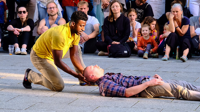 A man is kneeling and holding a person's head who is lying on the ground, surrounded by an audience of various ages sitting on the pavement.