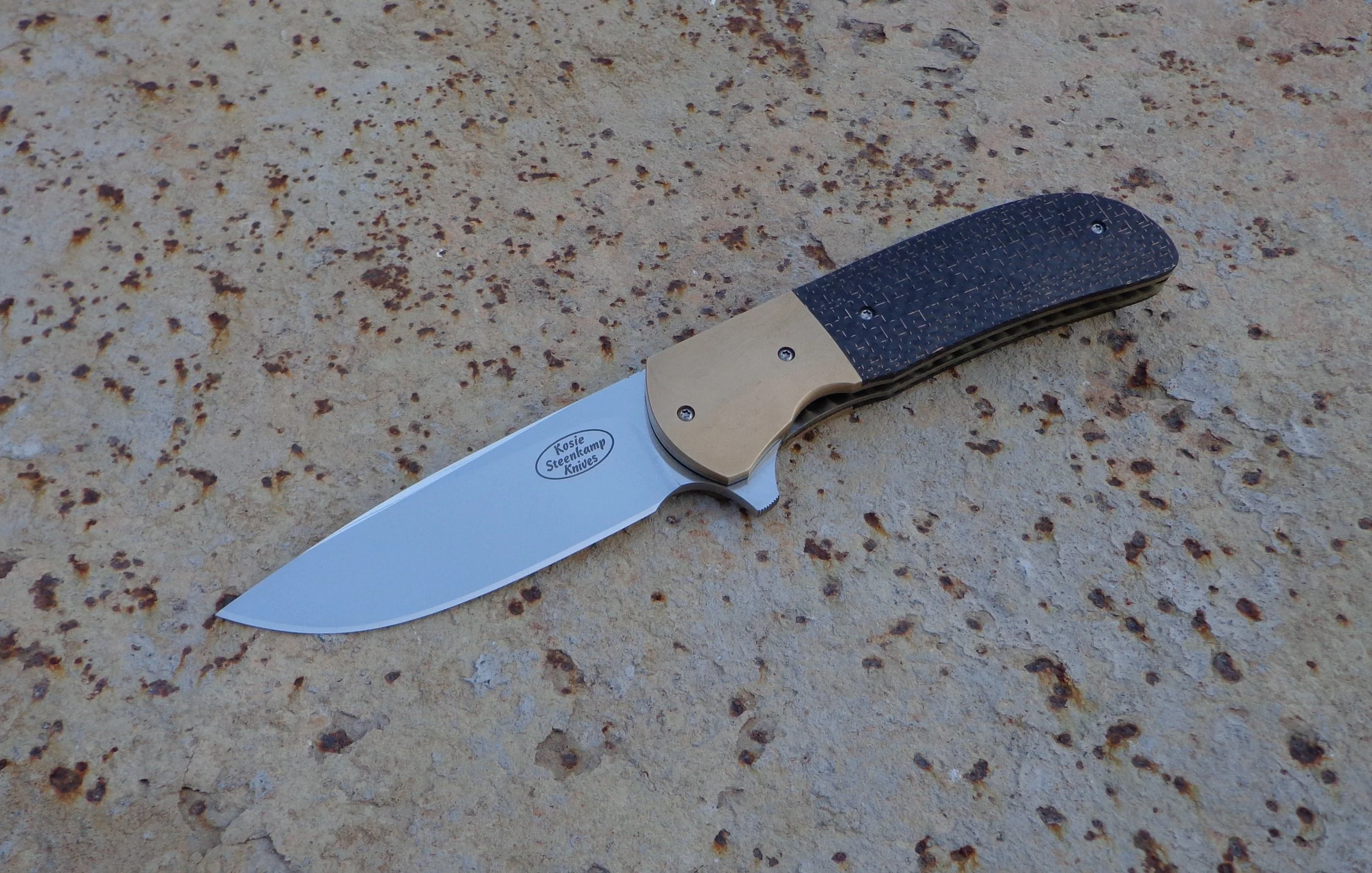 Knife: Piuma Flipper. Blade: Böhler N690 Stainless Steel. Bolsters: Bronze. Handle scales: LS Carbon Fiber. Liners and pocket clip: Titanium, anodized golden.