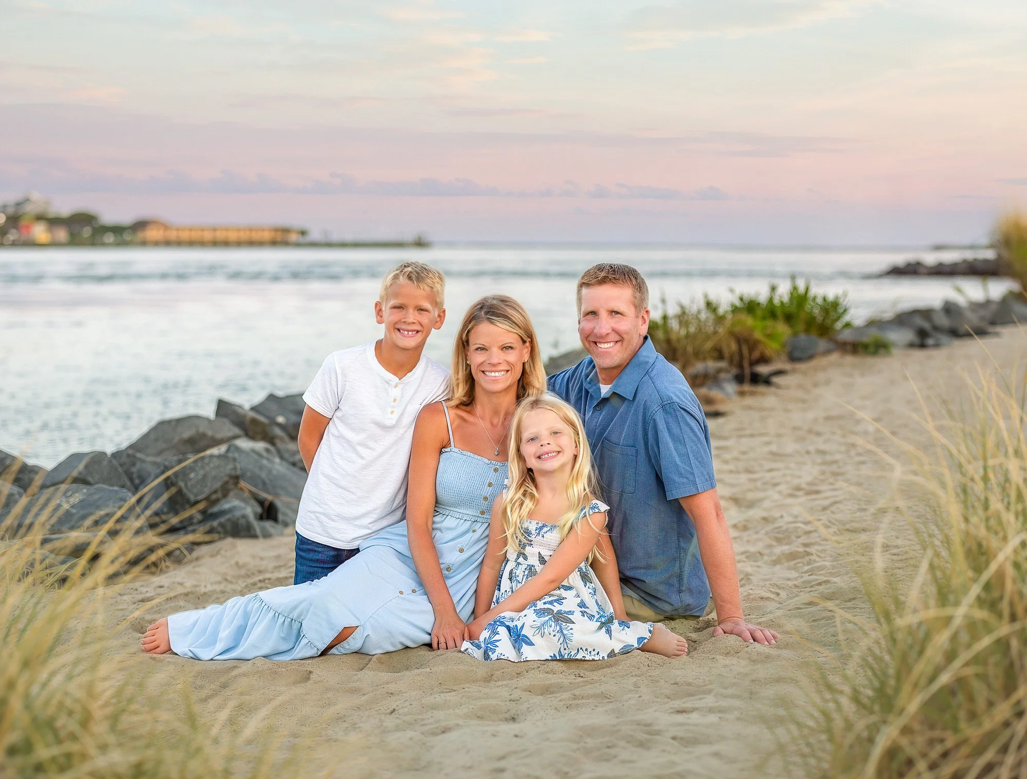 A family of five sitting on the sandy beach near rocks, smiling at the camera with the ocean and sky in the background.