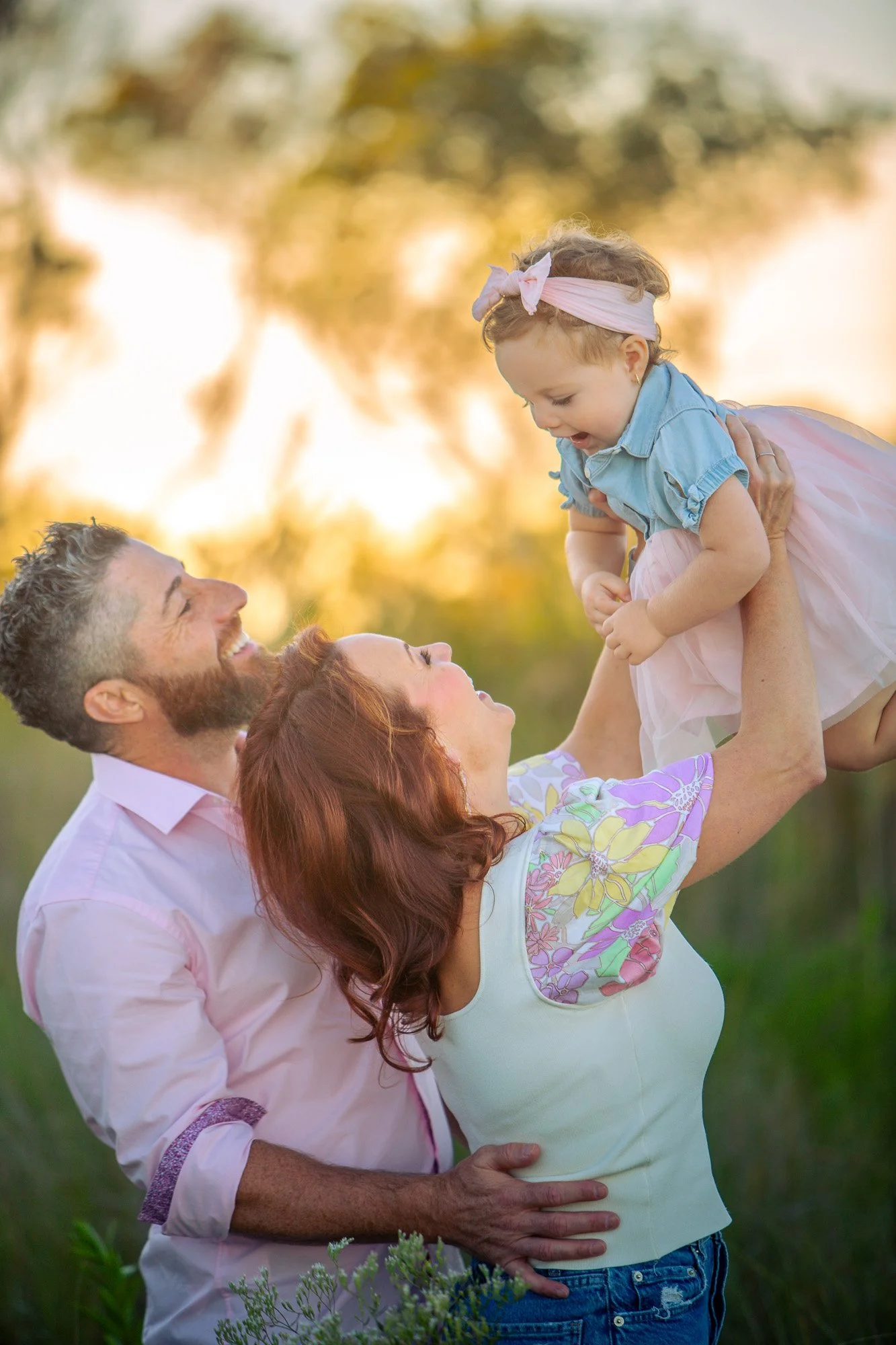 A family outdoors during sunset, with a man, a woman, and a young girl, the man lifting the girl in the air while the family smiles and enjoys the moment.