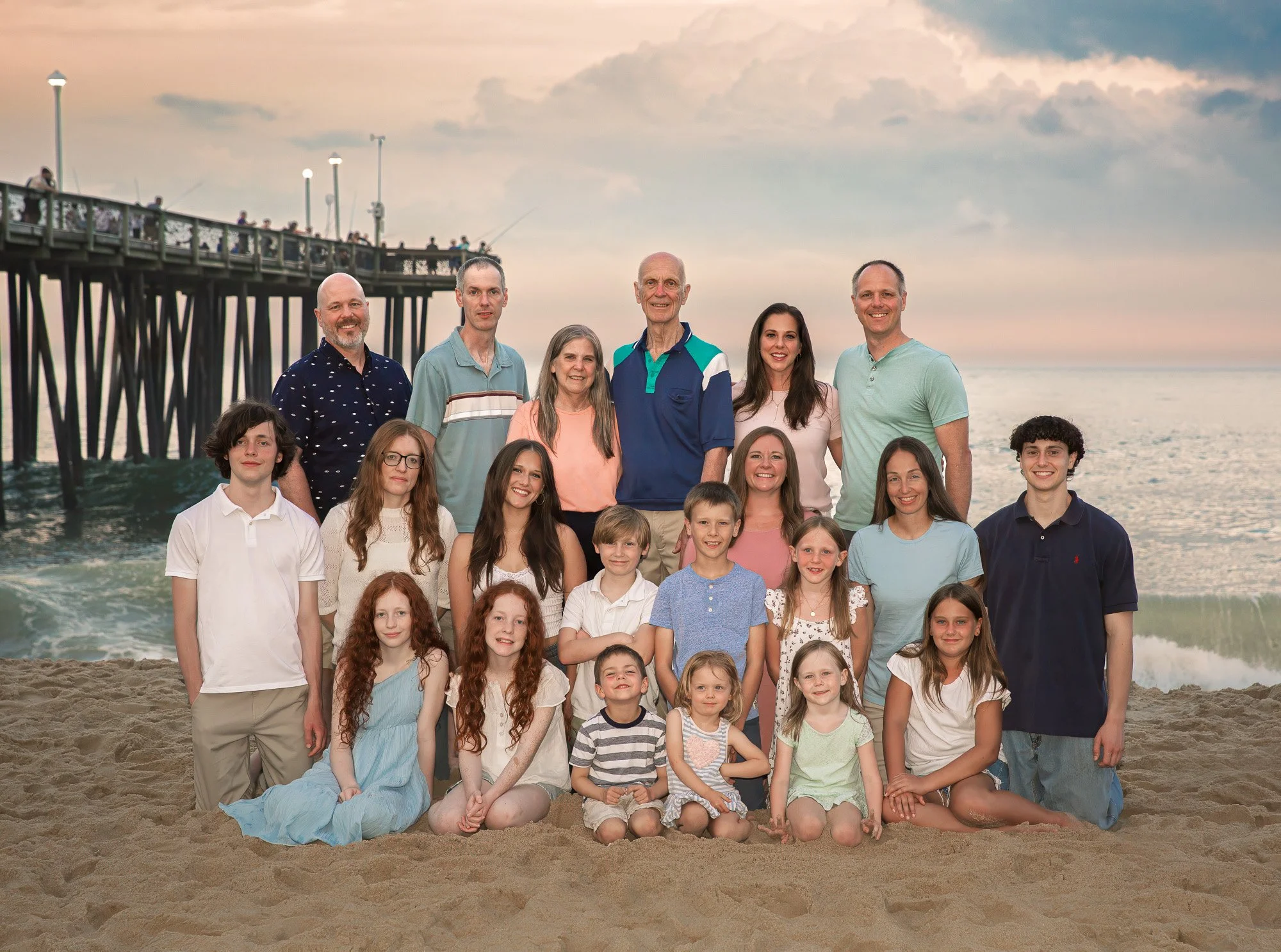 A large group of people, including adults and children, posing together on a sandy beach with a pier in the background during sunset or sunrise.