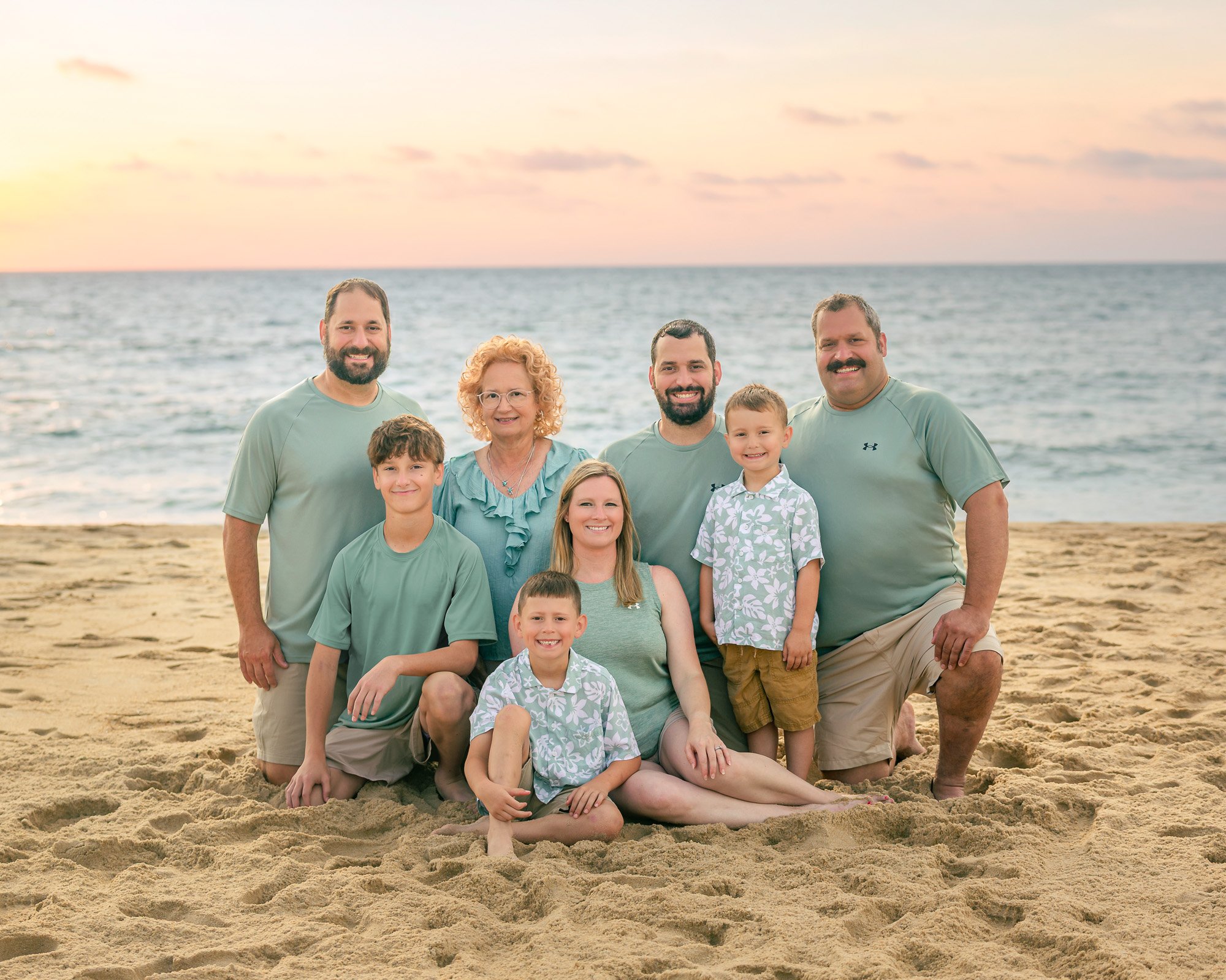 Family group photo on the beach at sunset, with sand in the foreground, the ocean in the background, and eight people smiling at the camera.