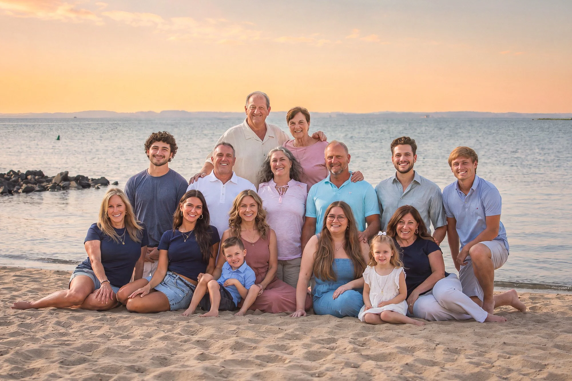 Group of people enjoying a beachside sunset, posing for a photo on the sand near the water, with a calm ocean and distant land in the background.