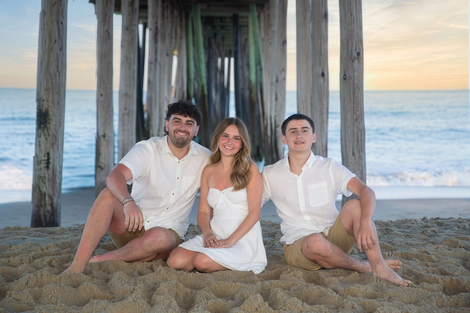 A group of three young adults, two men and one woman, sitting on the sandy beach under a pier during sunset. They are smiling and appear happy, wearing light-colored casual summer clothing.