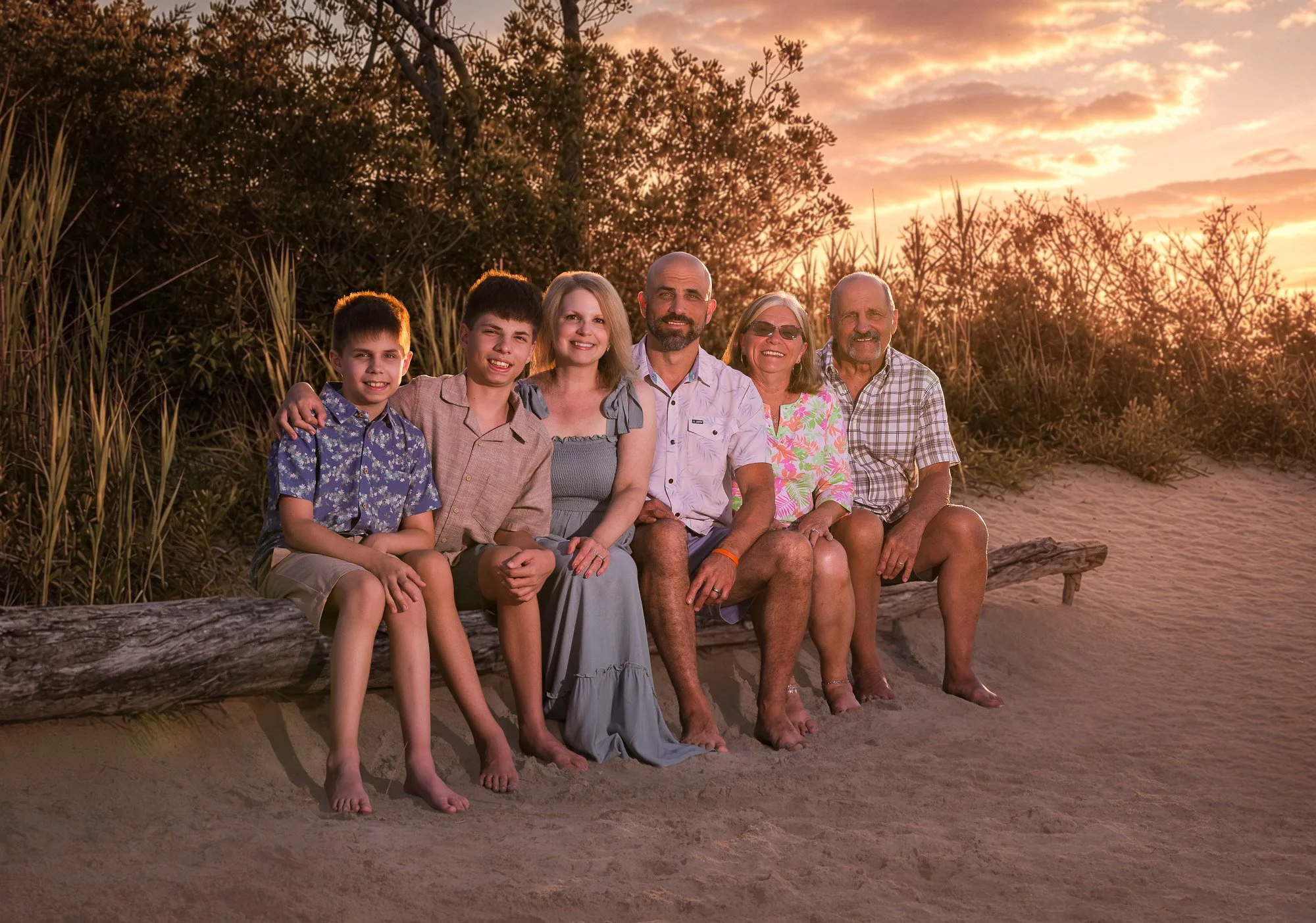 Family of seven sitting on a log on a beach during sunset, with trees and clouds in the background.