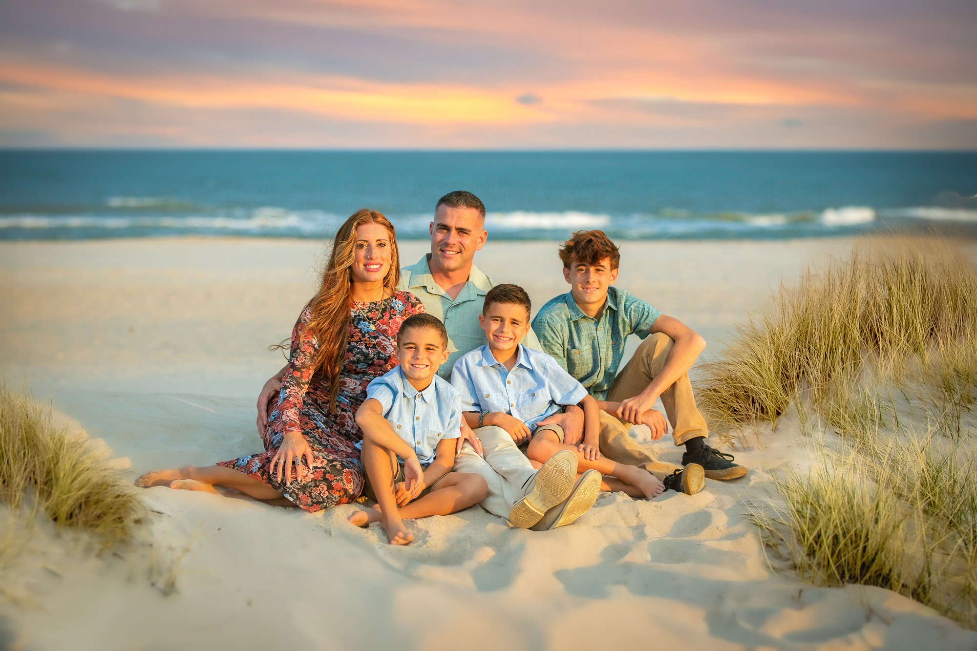 Family of six sitting on sandy beach with grass patches, smiling at the camera during sunset, with the ocean and colorful sky in the background.