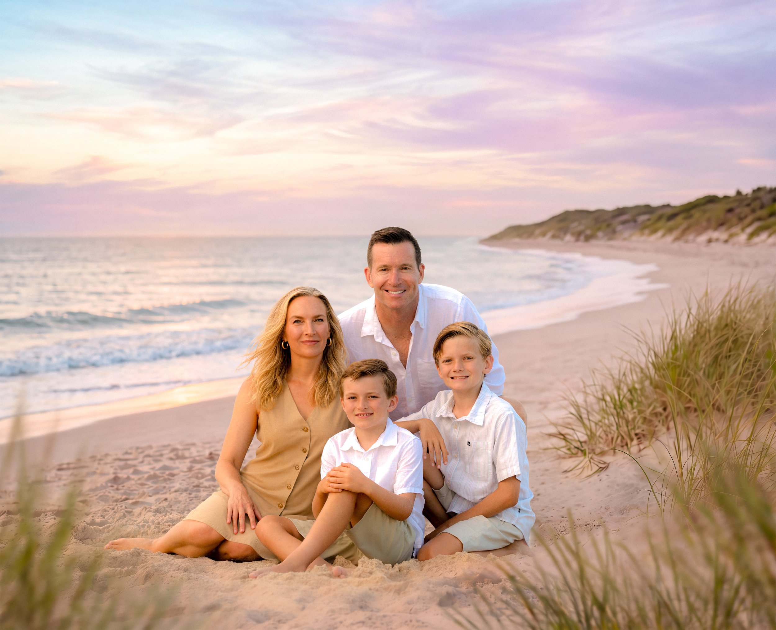 A family of five sitting on the beach during sunset, with the ocean and sand dunes in the background.