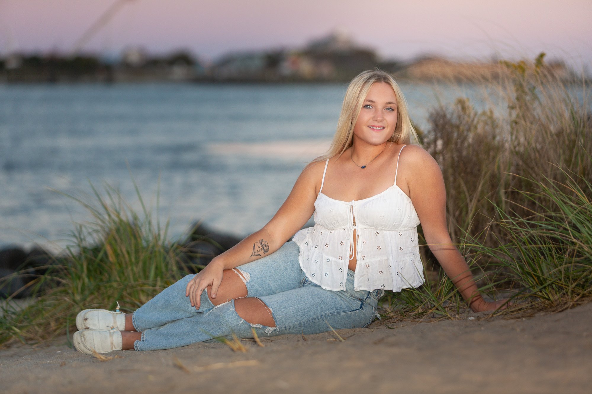A young woman with blond hair, smiling, wearing a white sleeveless top and ripped jeans, sitting on the sand near water with grass and distant buildings in the background during sunset.