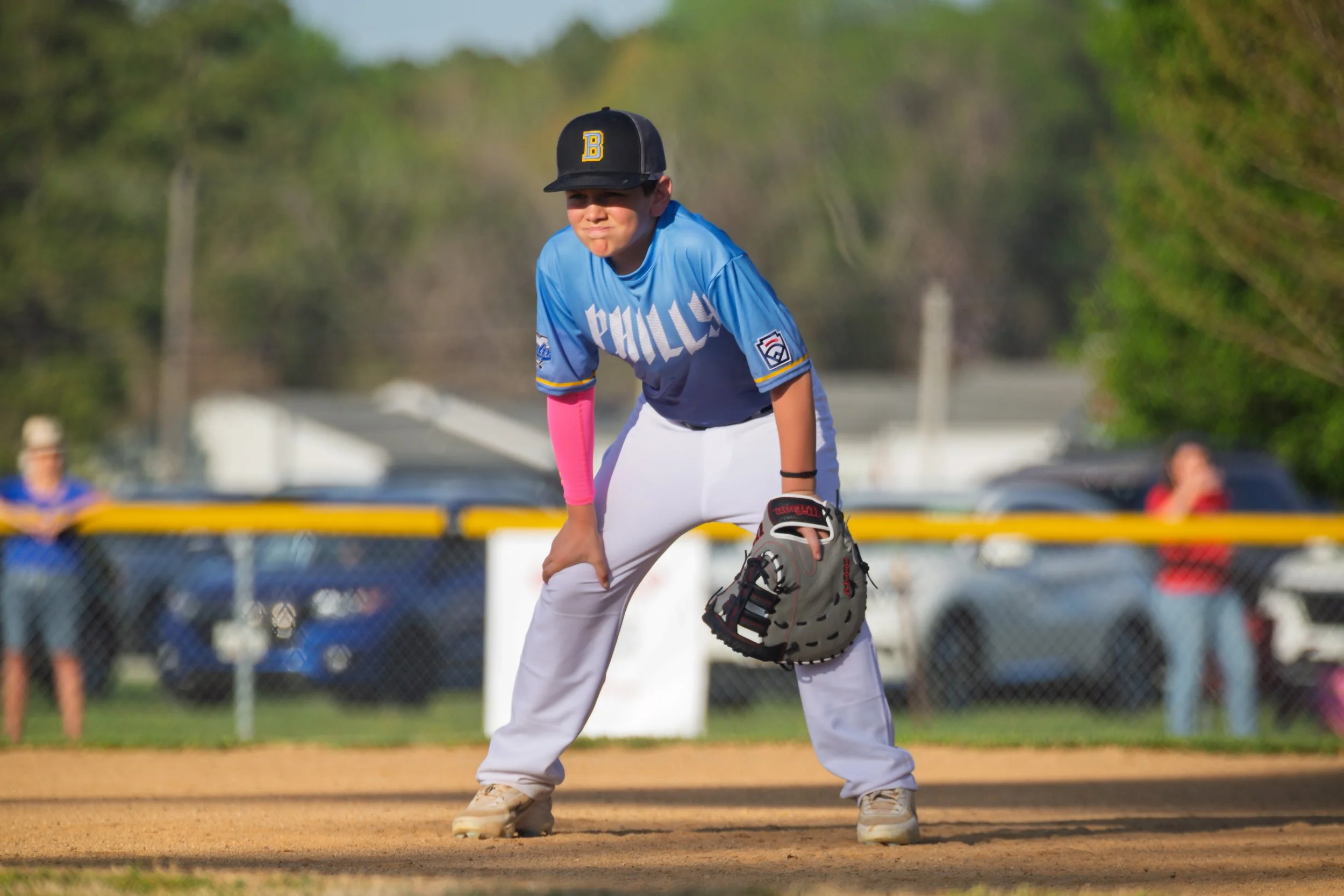 A young baseball player in a blue jersey and white pants crouches on the field, holding a glove, with a focused expression, near the baseball diamond with a chain-link fence and spectators in the background.