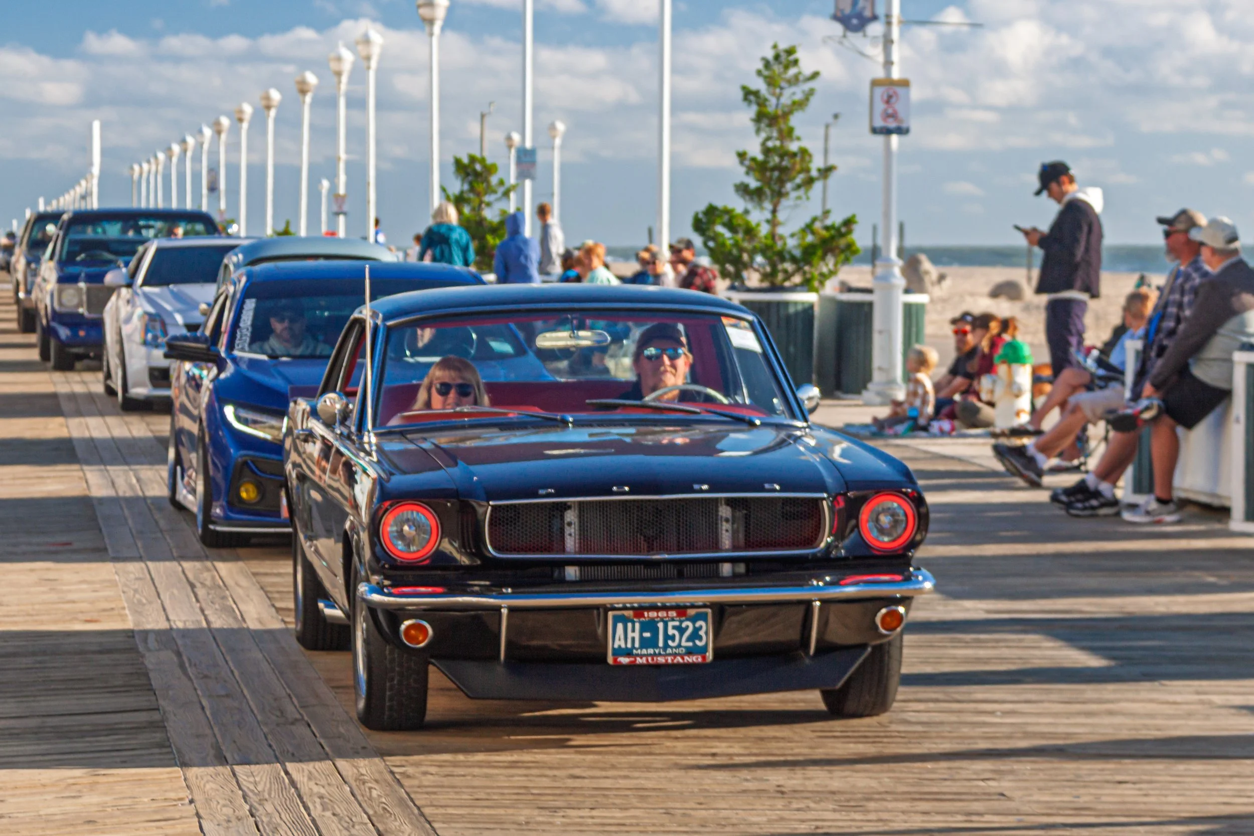 Classic cars, including a black Ford Mustang, are lined up on a wooden pier at a daytime car show. People are walking and sitting around, with some using their smartphones, near the beach under a partly cloudy sky.