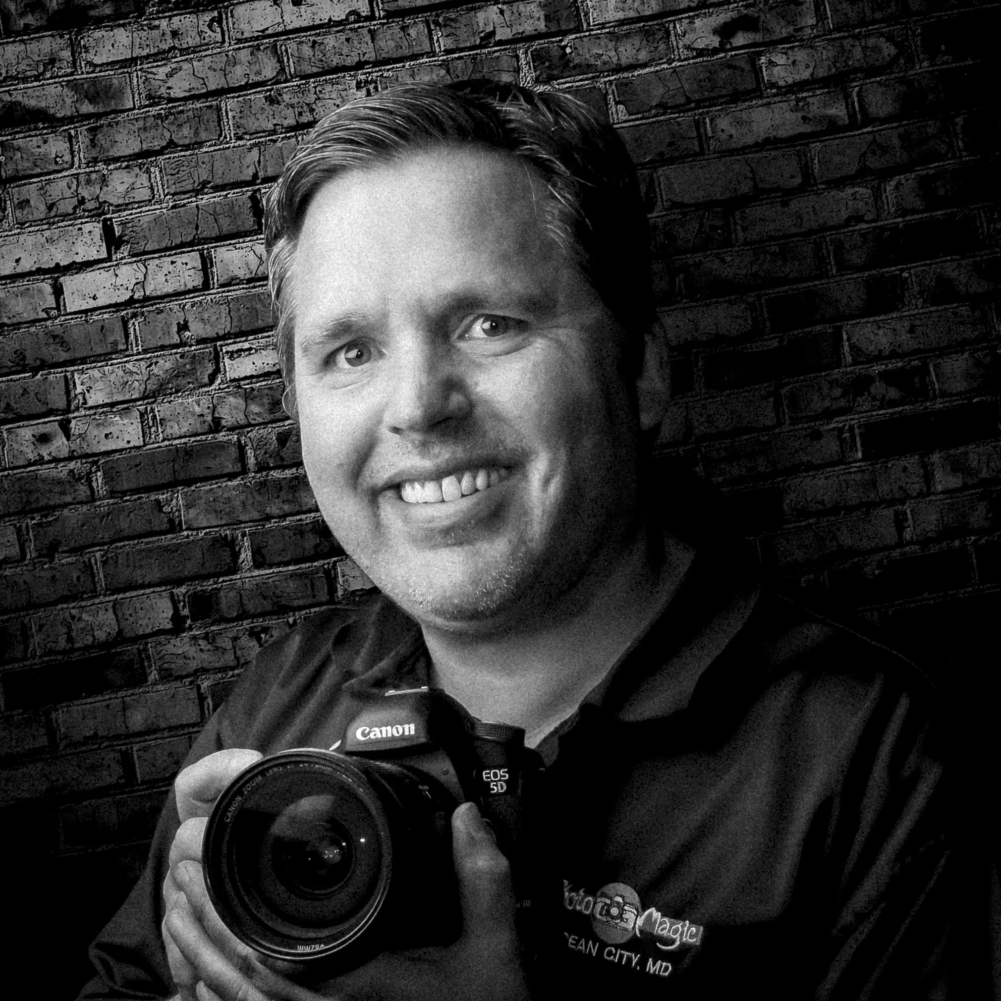 Black and white selfie of a man with short hair holding a Canon camera, standing in front of a brick wall.