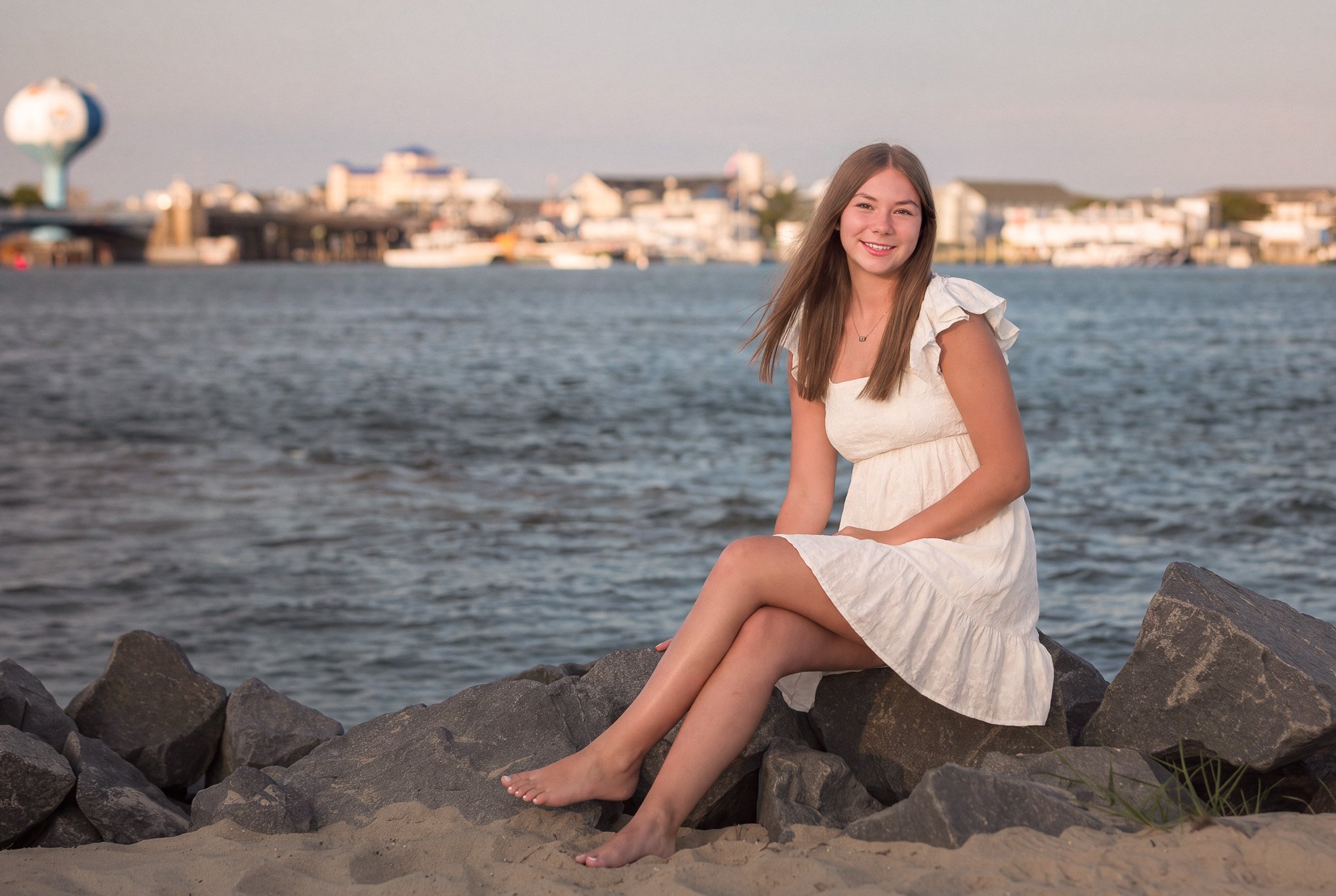 A young woman in a white dress sitting on rocks by the water, smiling at the camera, with a city skyline and a water tower in the background during late afternoon or early evening.