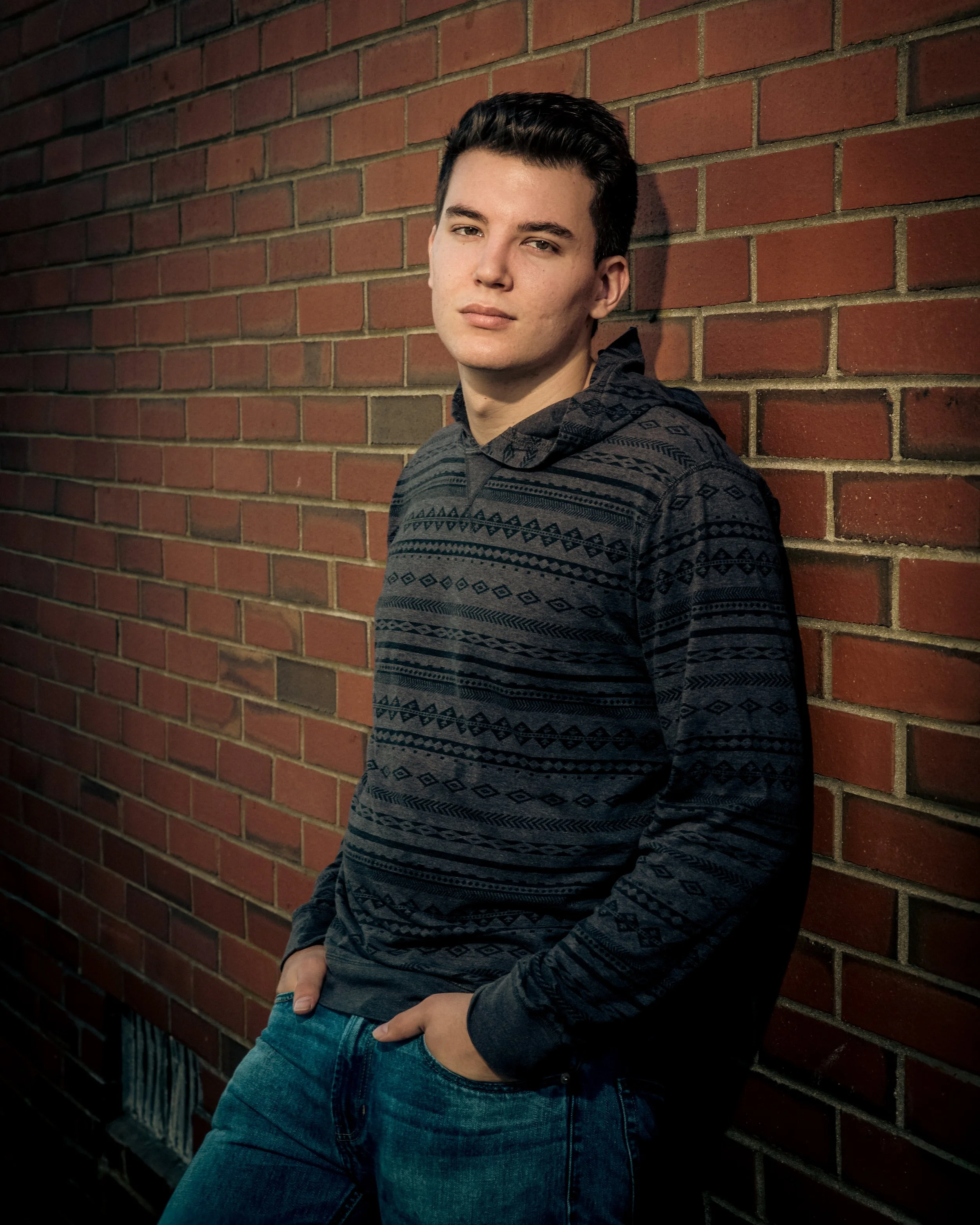 A young man with dark hair, wearing a patterned hoodie and jeans, leaning against a red brick wall with his hands in his pockets, looking at the camera with a serious expression.