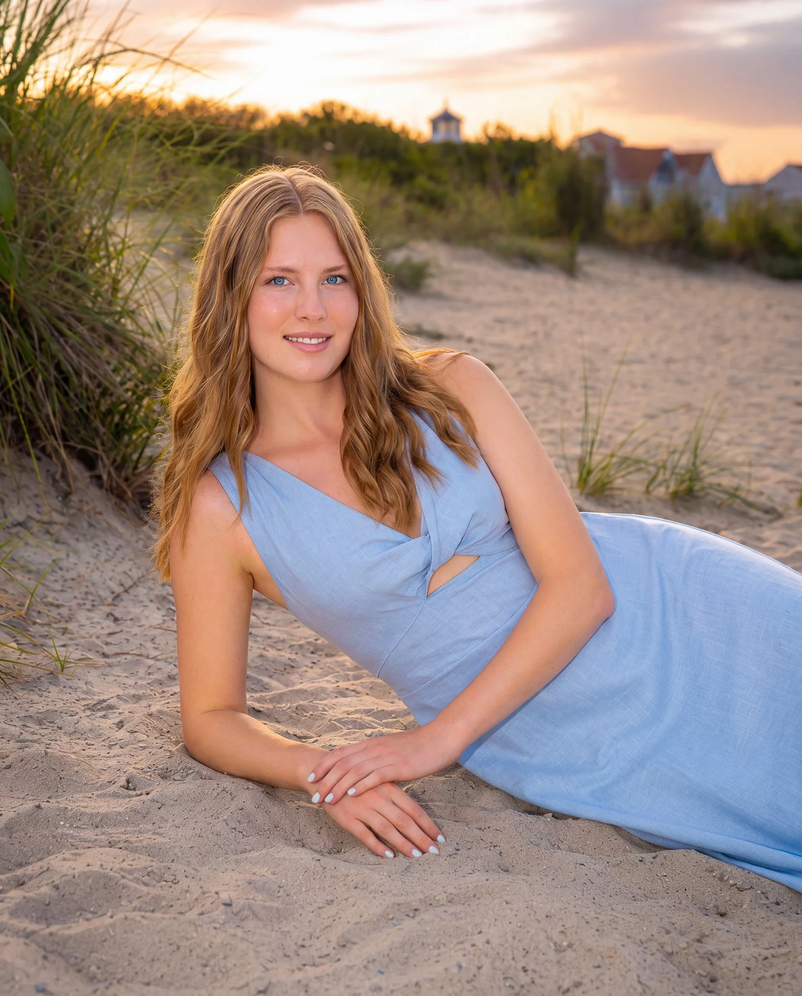 A woman with long blonde hair, blue eyes, and fair skin lying on the sandy ground near grass, wearing a light blue sleeveless dress, with a sunset sky and houses with red roofs in the background.