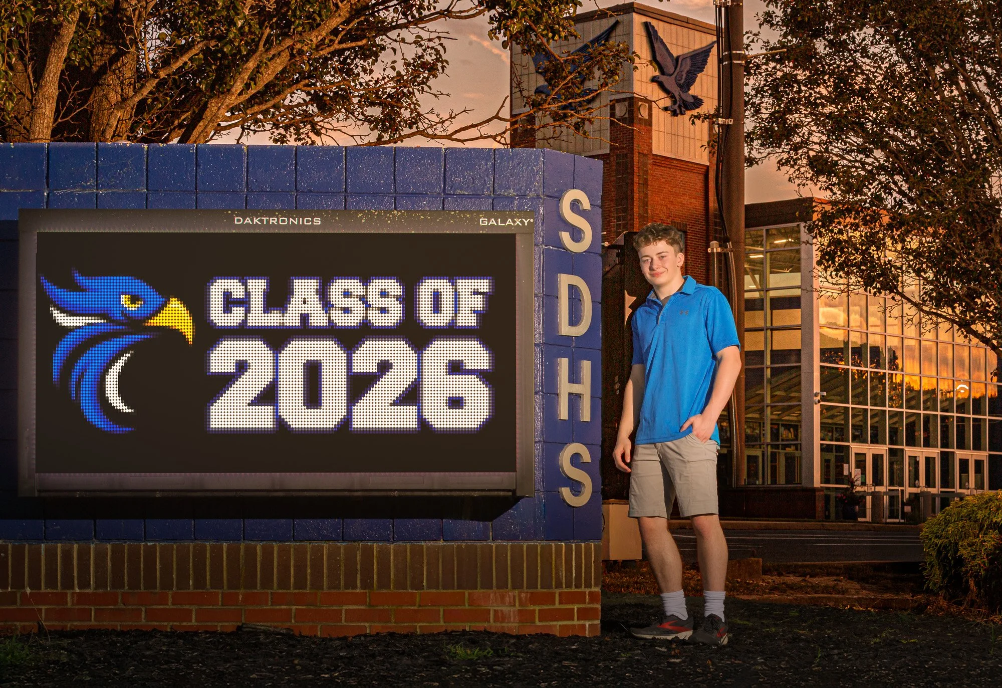 A teenage boy in a blue shirt and khaki shorts standing next to a sign that reads 'Class of 2026' with a blue and yellow bird logo. The sign is outside a school building with trees and a brick wall, during sunset.