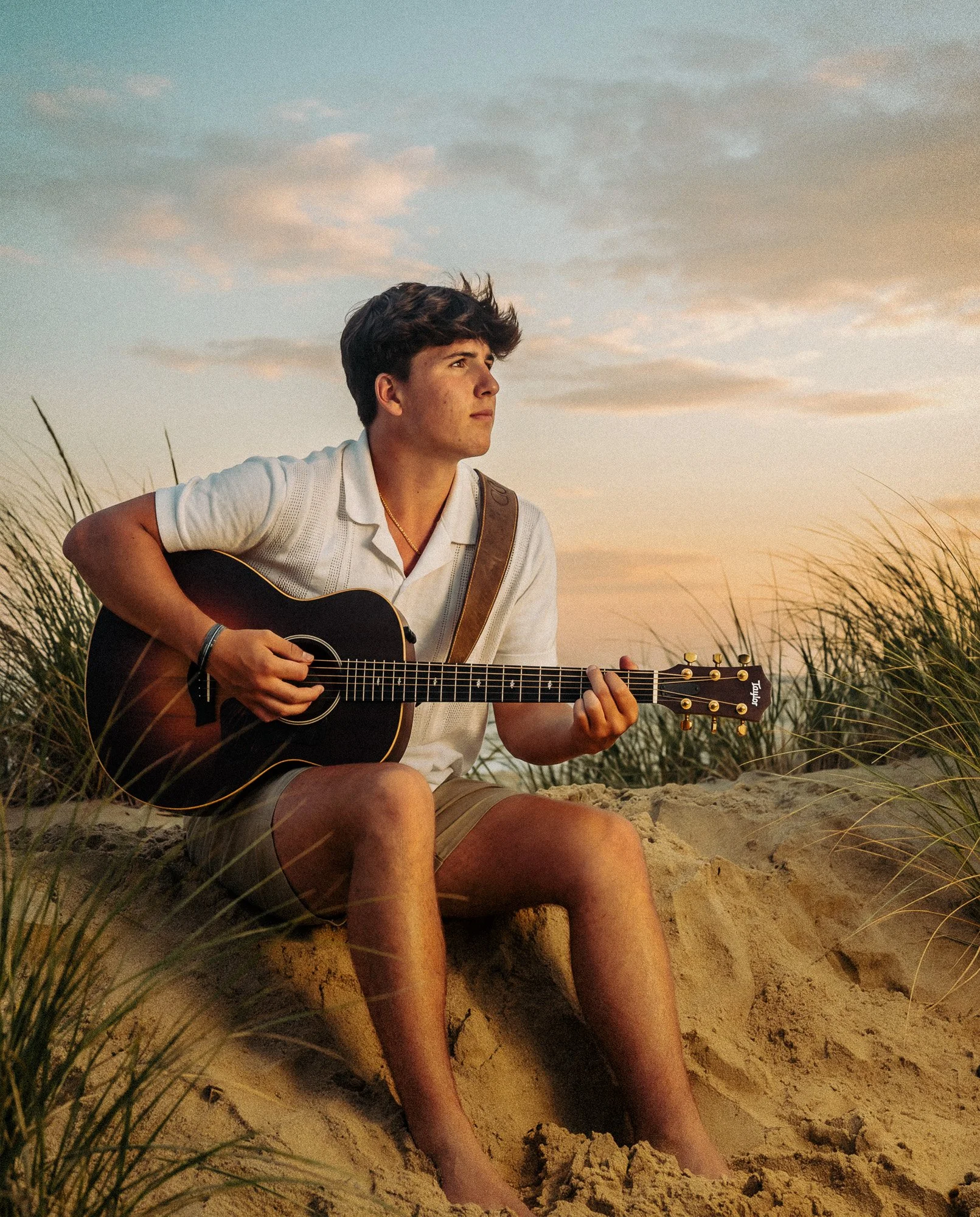 Young man sitting on sandy beach, playing guitar during sunset, surrounded by grass, with a cloudy sky.