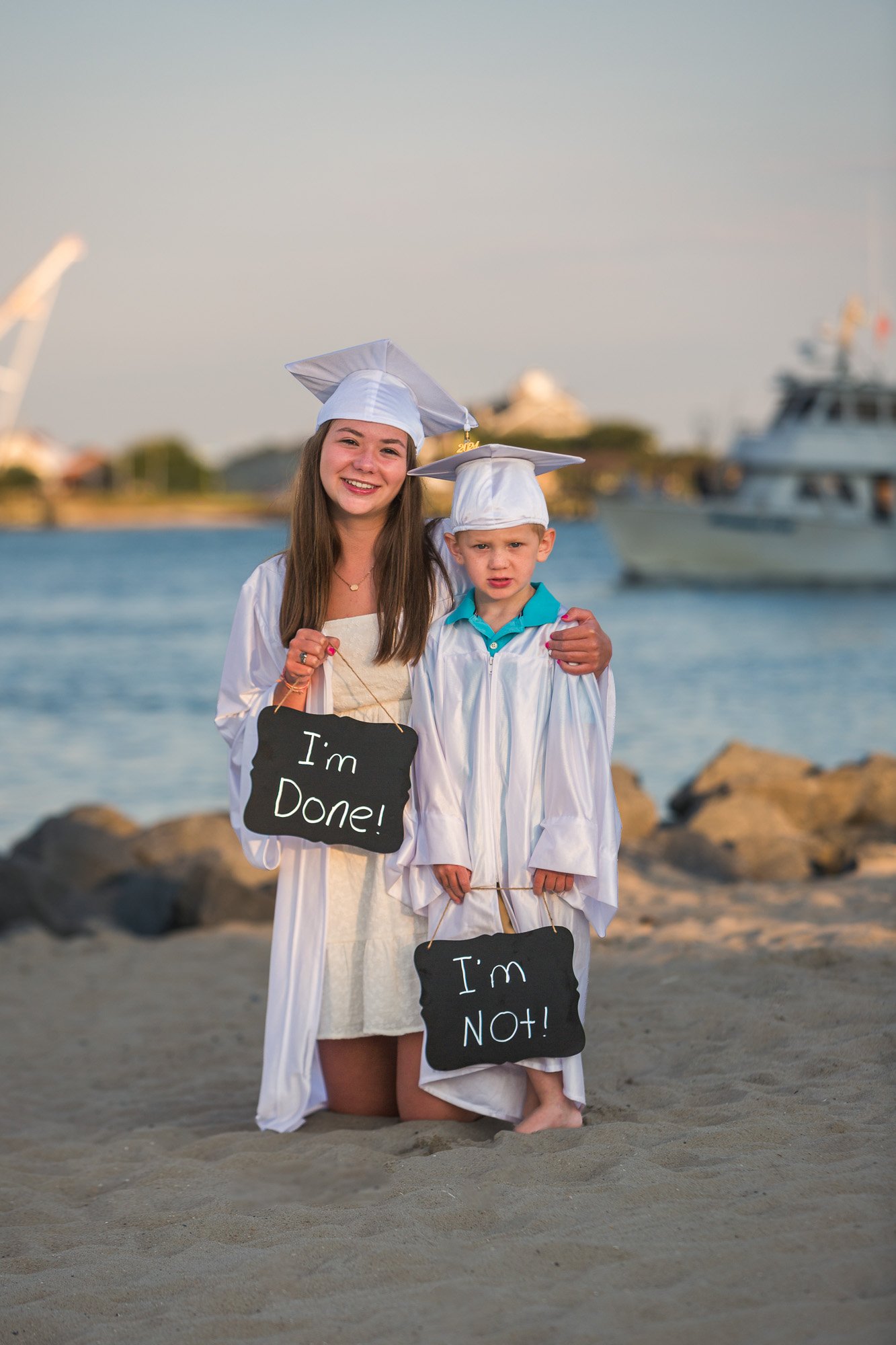 A young woman and a young boy in graduation caps and gowns standing on a beach with boats and a marina in the background. They are holding chalkboard signs: the woman’s sign says "I'm Done!" and the boy’s sign says "I'm NOT!".