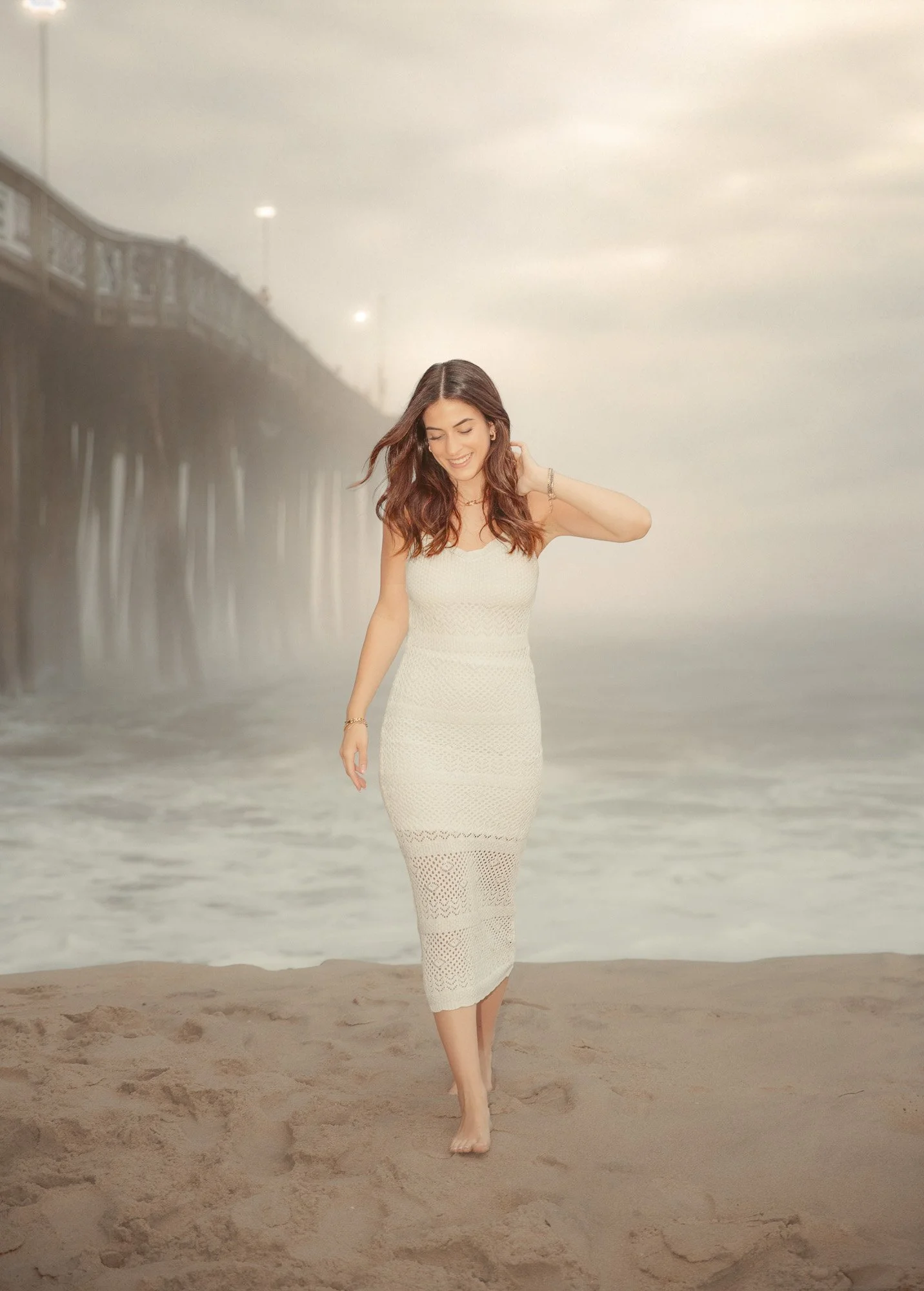 A woman with long brown hair wearing a white lace dress walking on the beach near the ocean with a pier and cloudy sky in the background.
