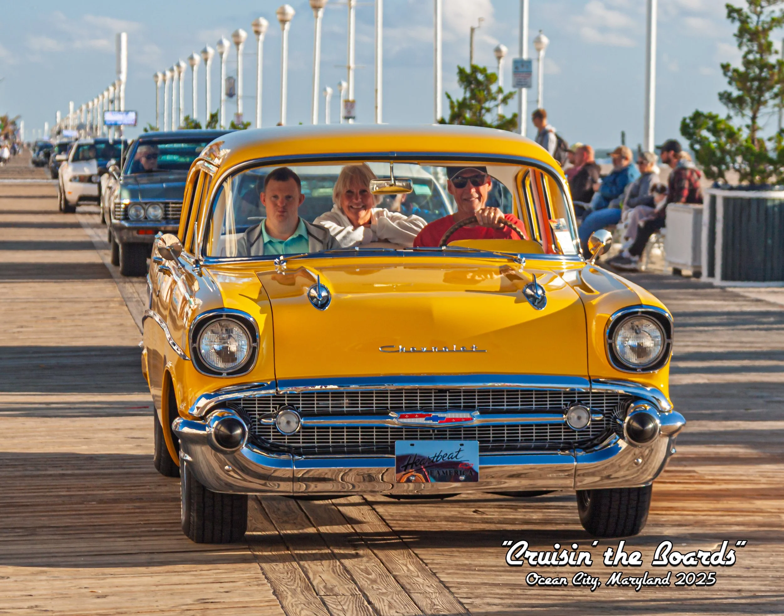 A yellow vintage Chevrolet car with three people inside, driving on a wooden boardwalk with cars parked and people sitting on benches along the side, in Ocean City, Maryland, 2025.