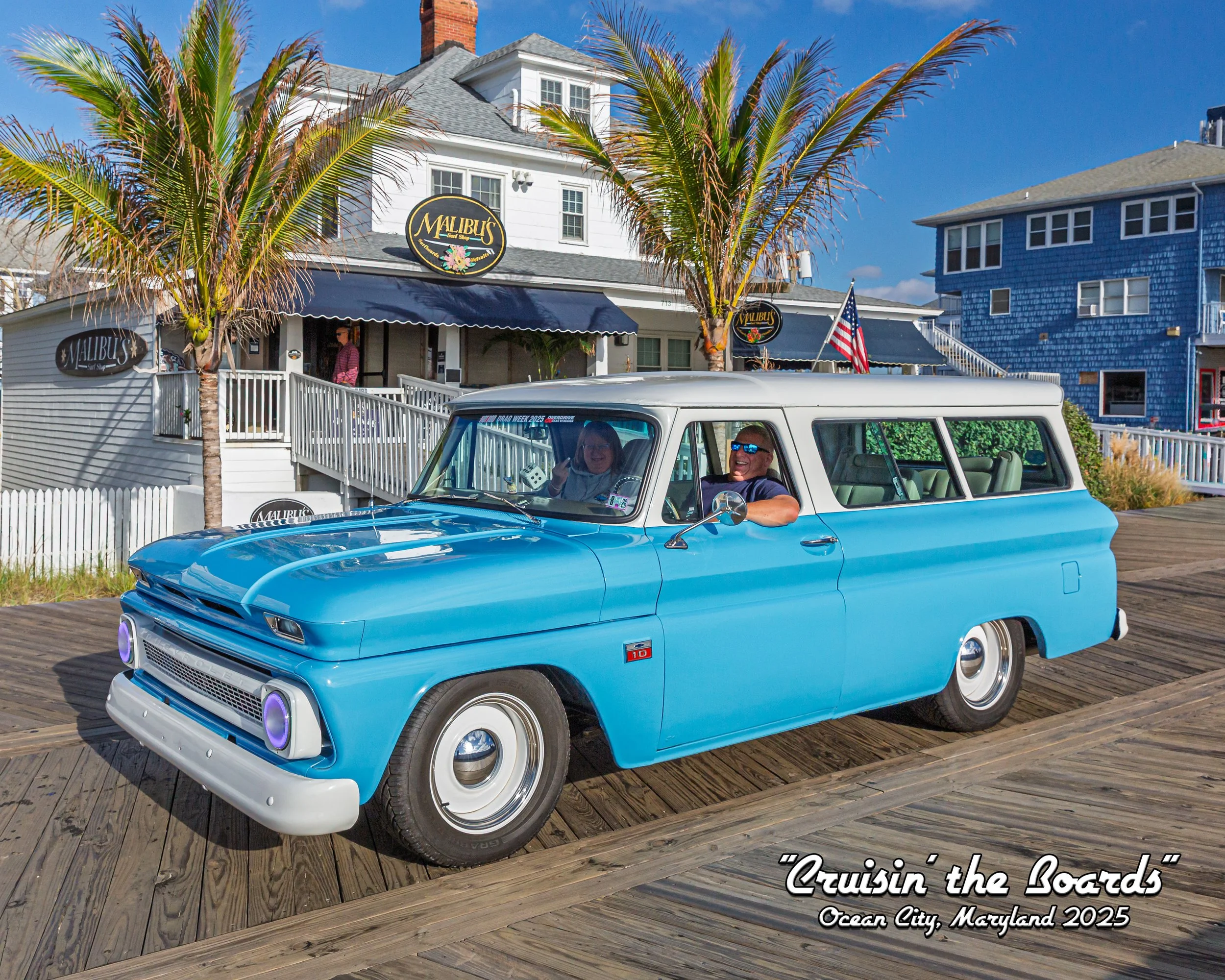 A vintage blue and white excursion vehicle with two passengers smiling and waving, in front of a white beach house with palm trees and a sign for Malibus Surf Shop, at Ocean City, Maryland in 2025.