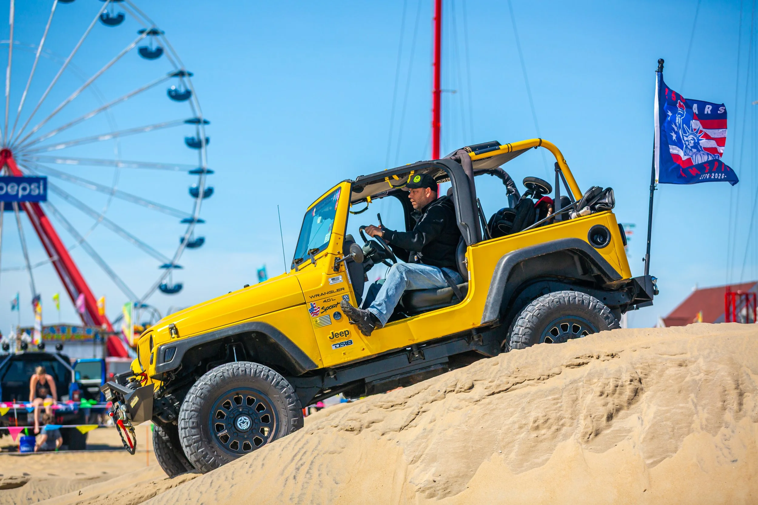 A man in a black jacket and cap sitting in a yellow Jeep Wrangler off-road vehicle on a sandy hill at a beach amusement park, with a Ferris wheel and colorful flags in the background.