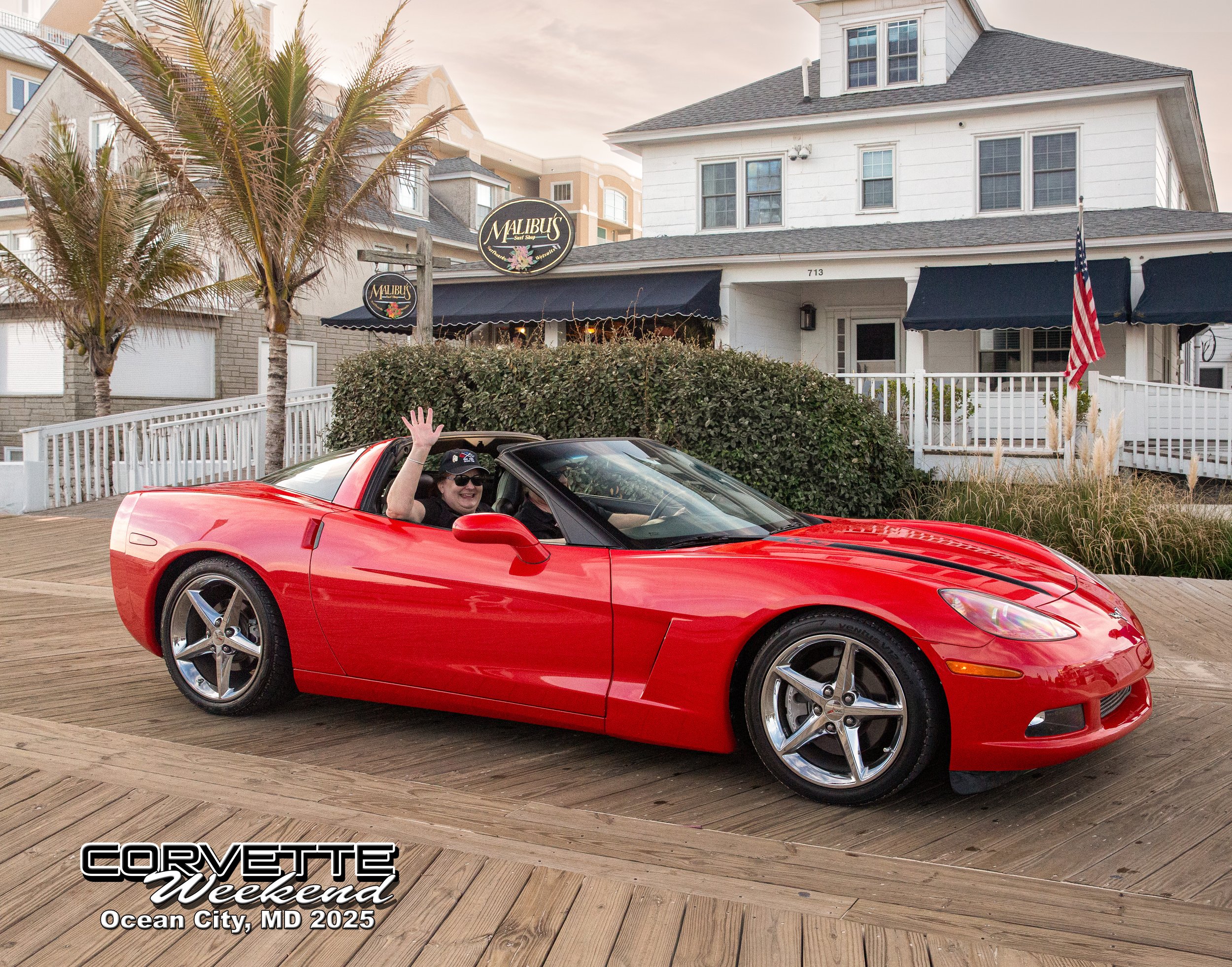 A smiling woman waving from the driver's seat of a red Chevrolet Corvette sports car parked on a wooden deck in front of a white house with black awnings, a palm tree, and American flag. The image has text overlay that says "Corvette Weekend Ocean City, MD 2025."