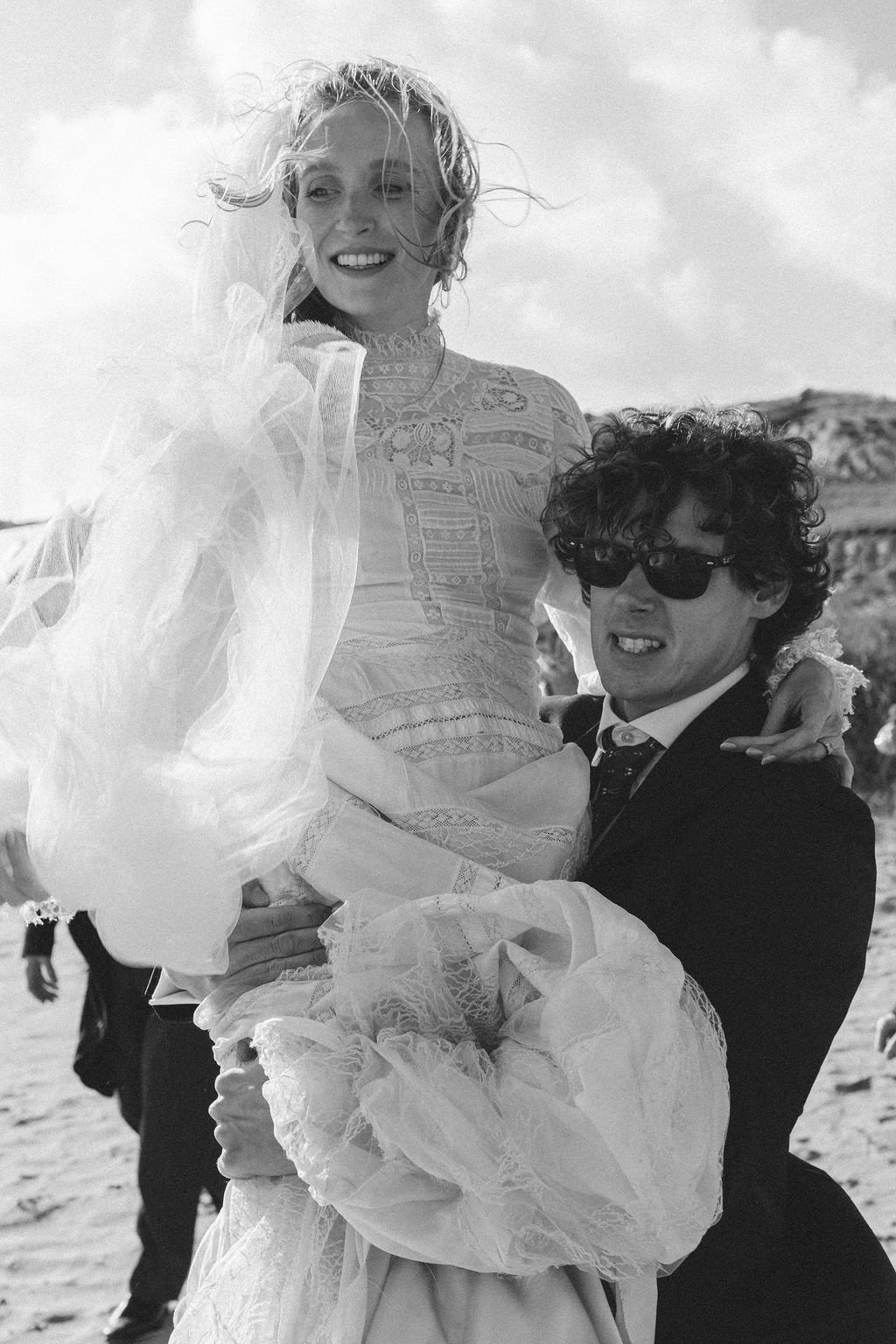 A woman in a wedding dress being held up by a man in a suit with sunglasses at the beach.