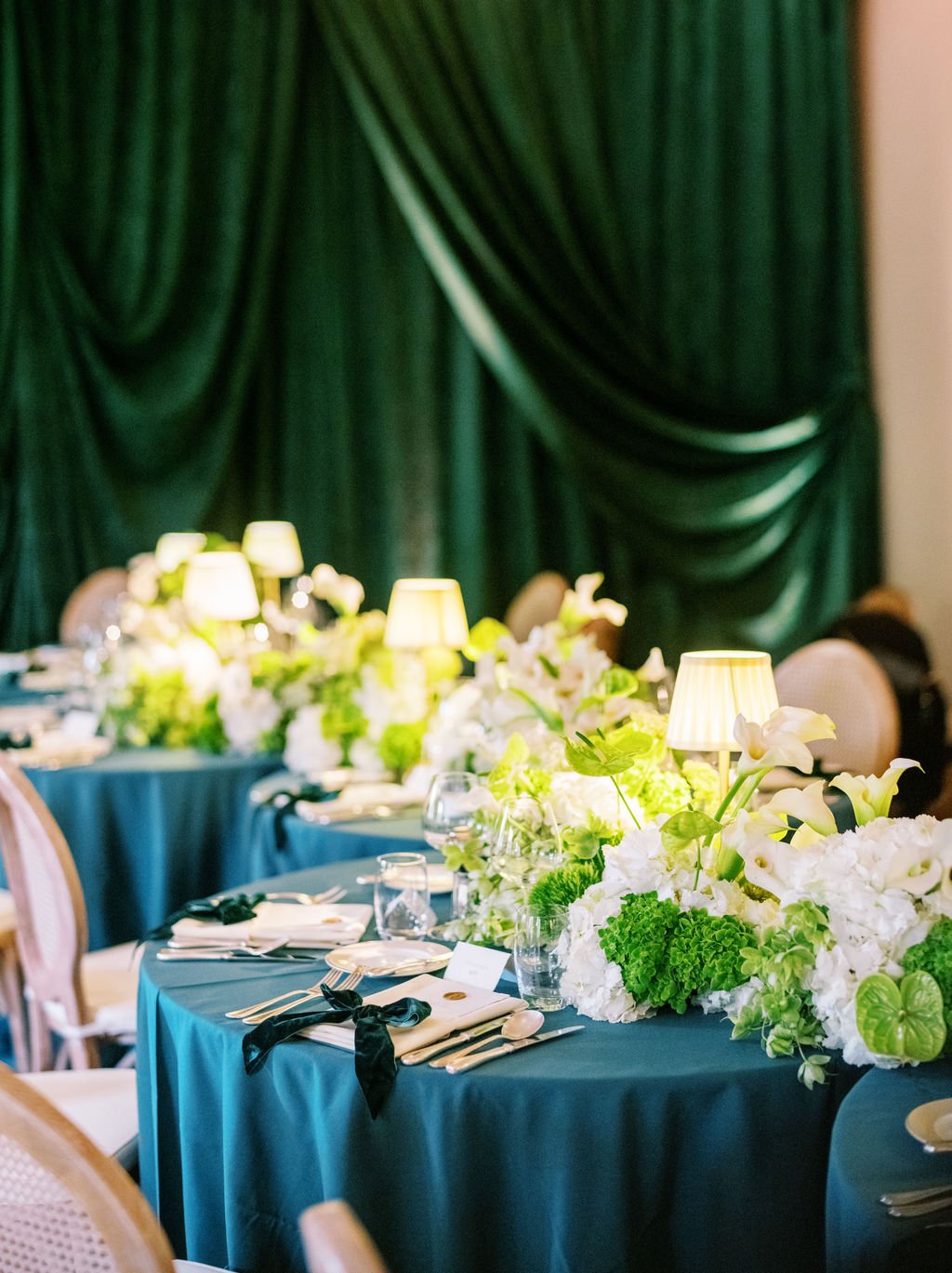 Elegantly set dining table with dark blue tablecloth, white floral centerpiece, small table lamps, glassware, silverware, and place settings, with green curtains in the background.