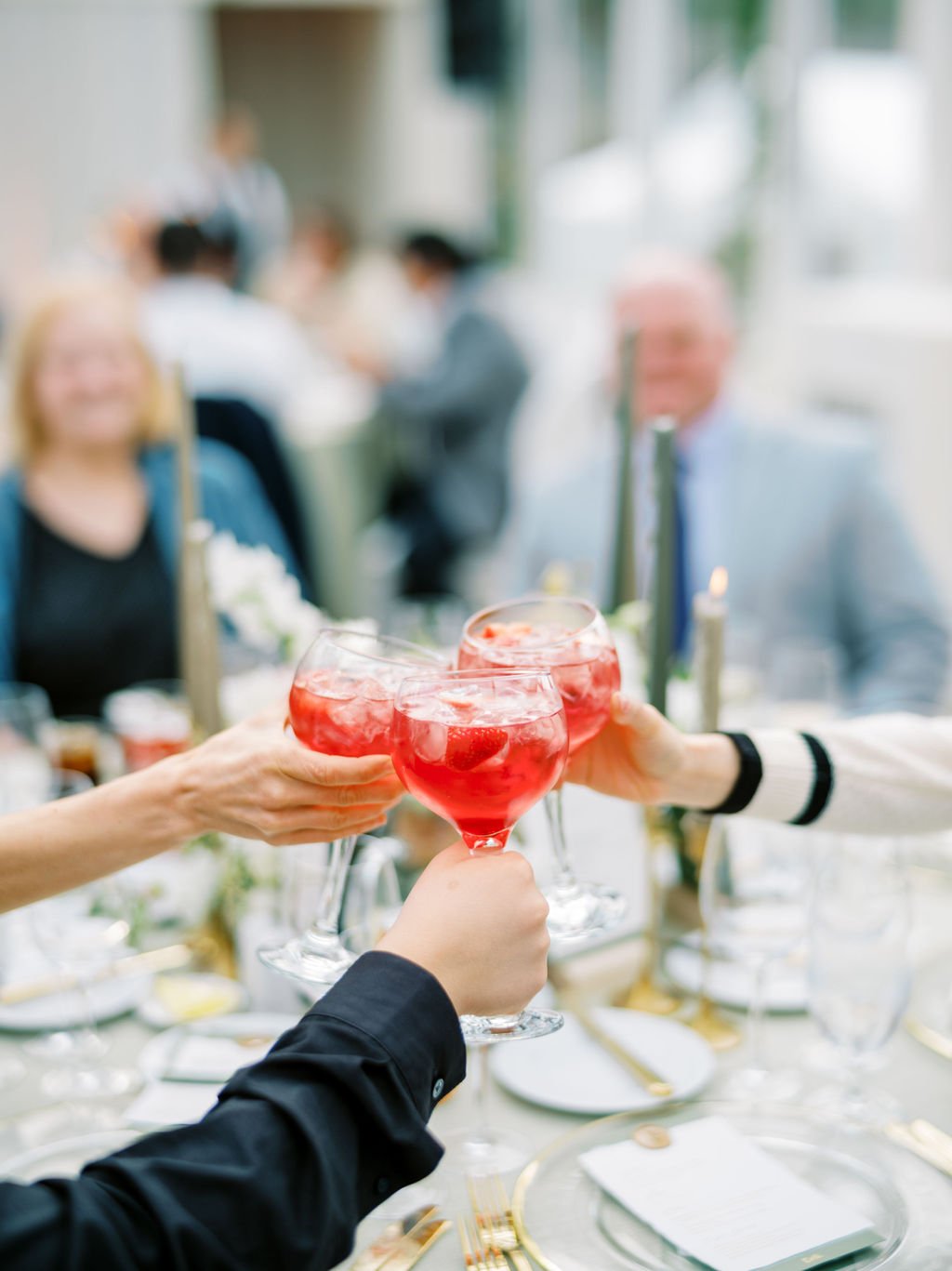 People toasting with red drinks at a social gathering, with blurred background of other guests and table settings.