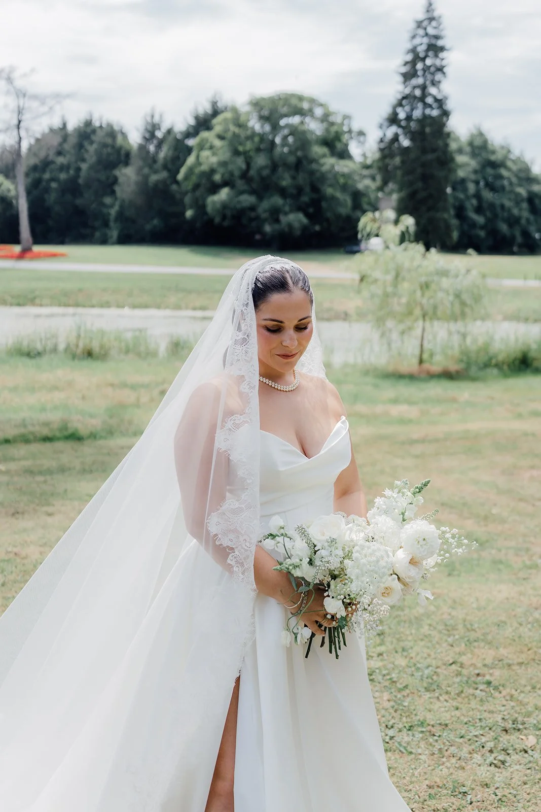 Bride standing outdoors in a white wedding gown with a long veil, holding a bouquet of white flowers, on a grassy area with trees and cloudy sky in the background.