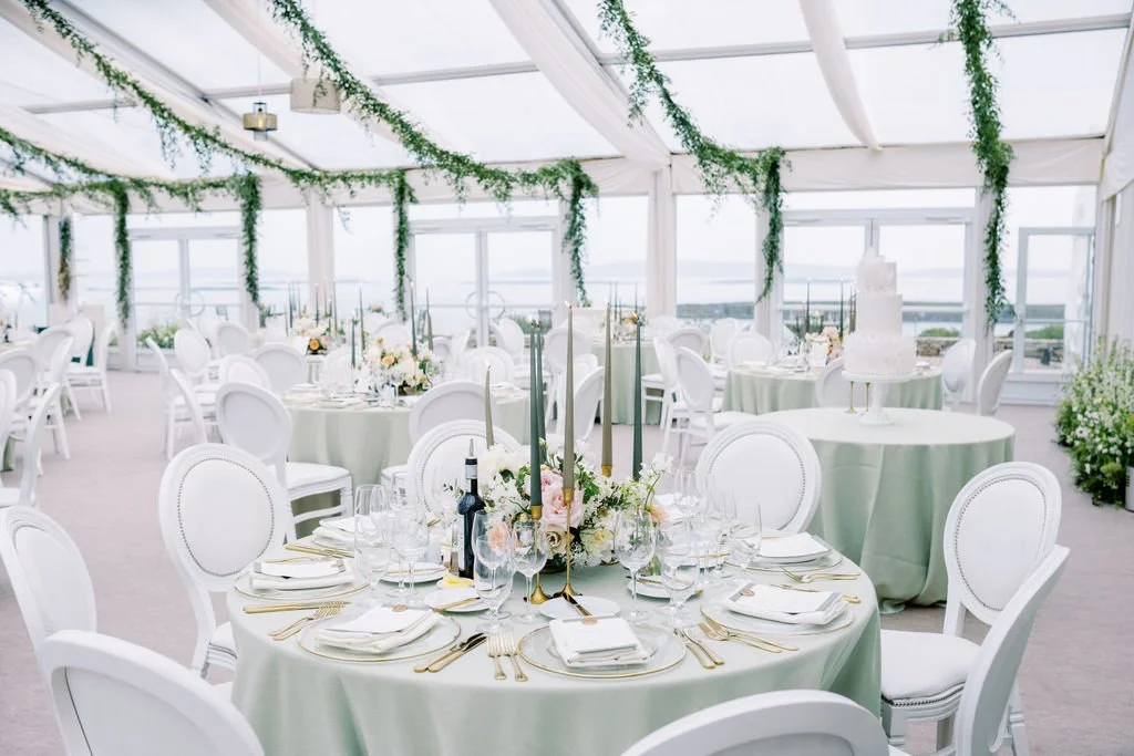 Elegant wedding reception area with round tables covered in light green tablecloths, decorated with floral centerpieces, candles, and gold cutlery. The room features large windows, white chairs, and green hanging vines.