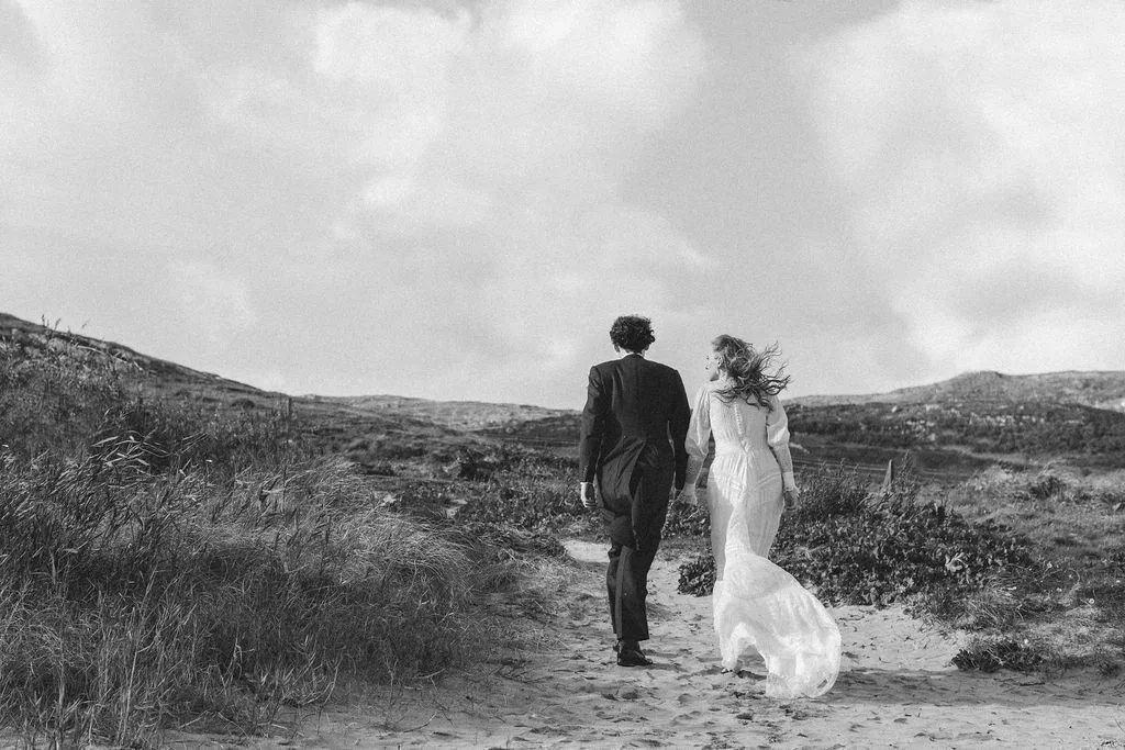 A couple dressed in wedding attire walking hand in hand along a sandy path in an open landscape with hills and cloudy sky.