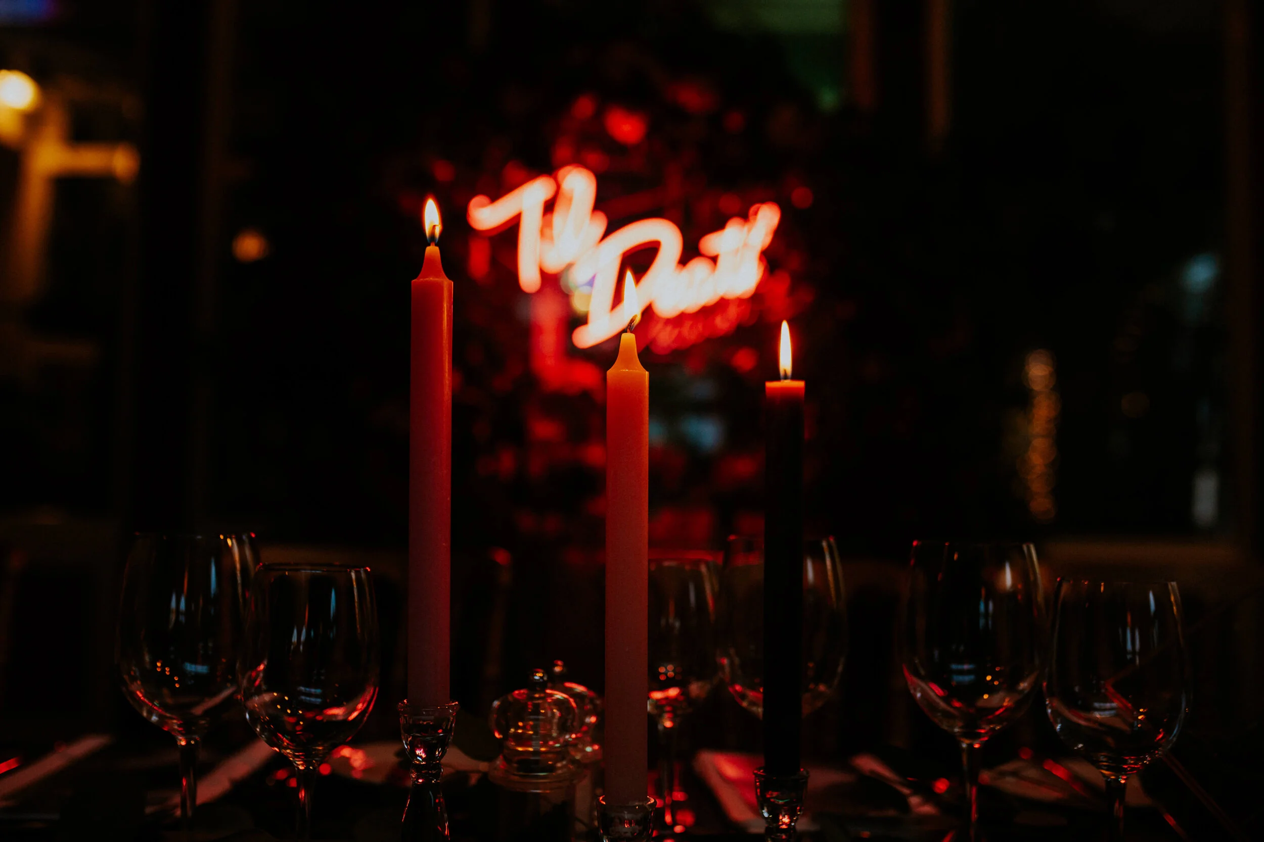 A dimly lit restaurant table with three lit candles in the foreground and empty wine glasses. In the background, a red neon sign reading 'The Duck' is visible.
