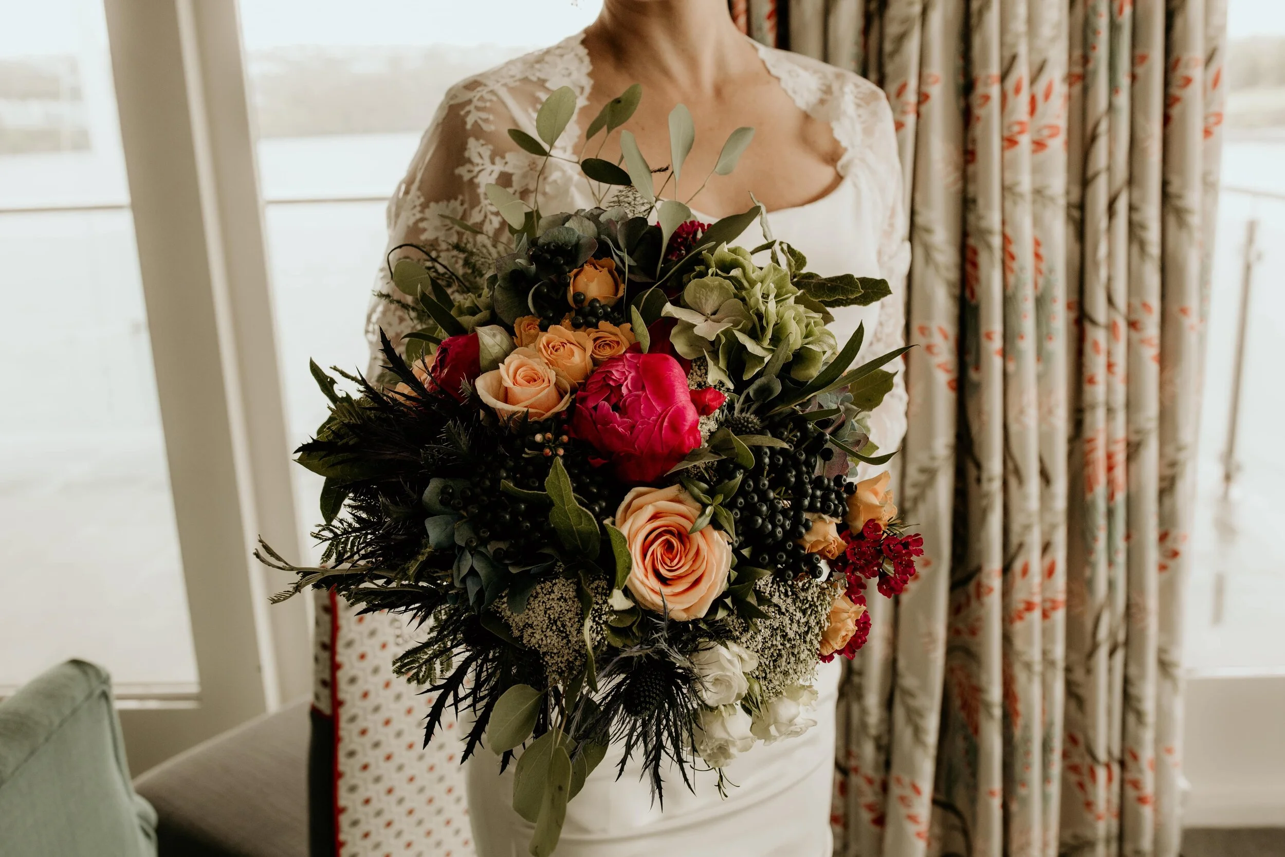 A bride holding a bouquet of various flowers including roses, peonies, and greenery, standing indoors near a window with floral curtains.