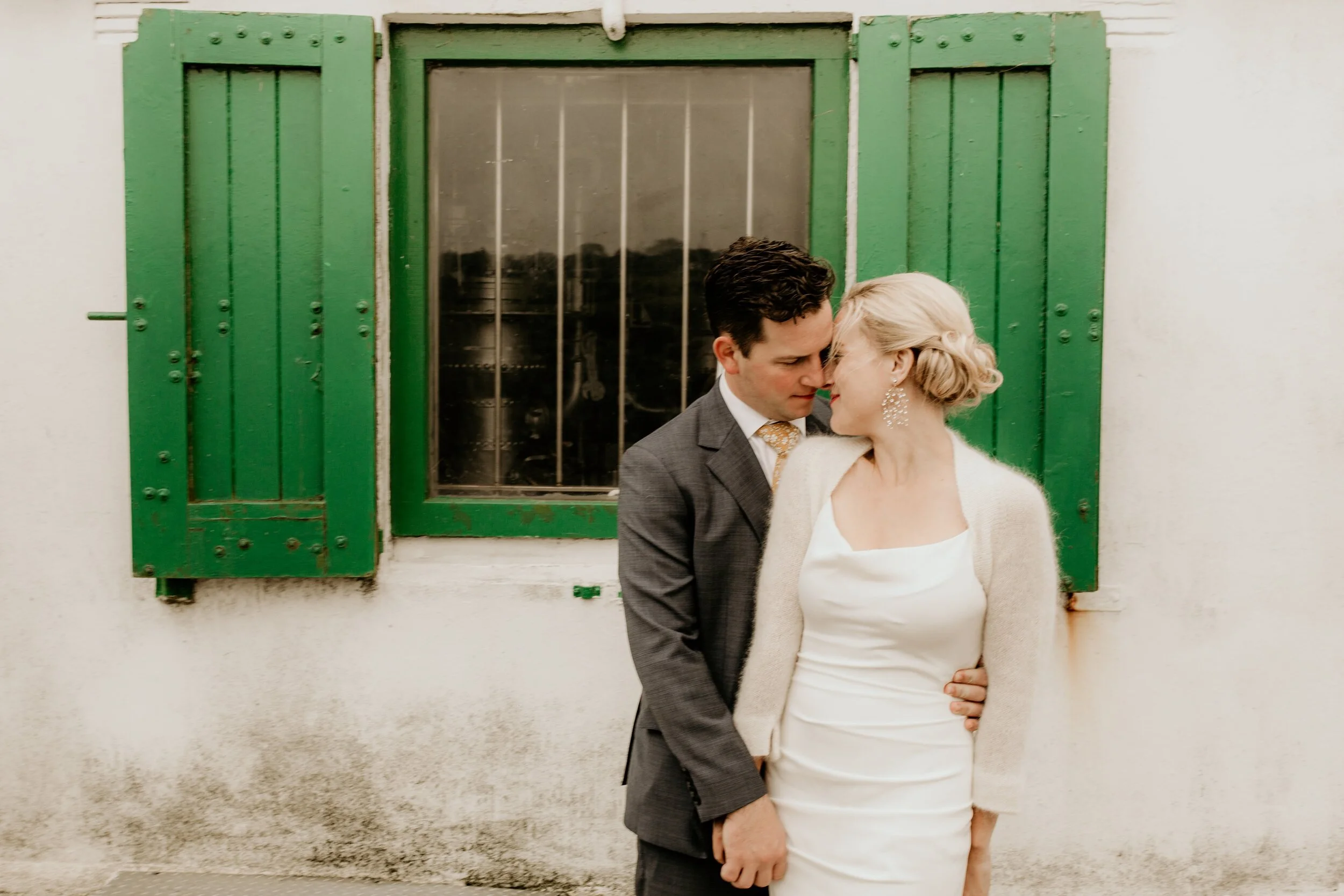 A couple dressed in wedding attire standing close together in front of a white wall with a window with green shutters. They are leaning their foreheads and noses together.