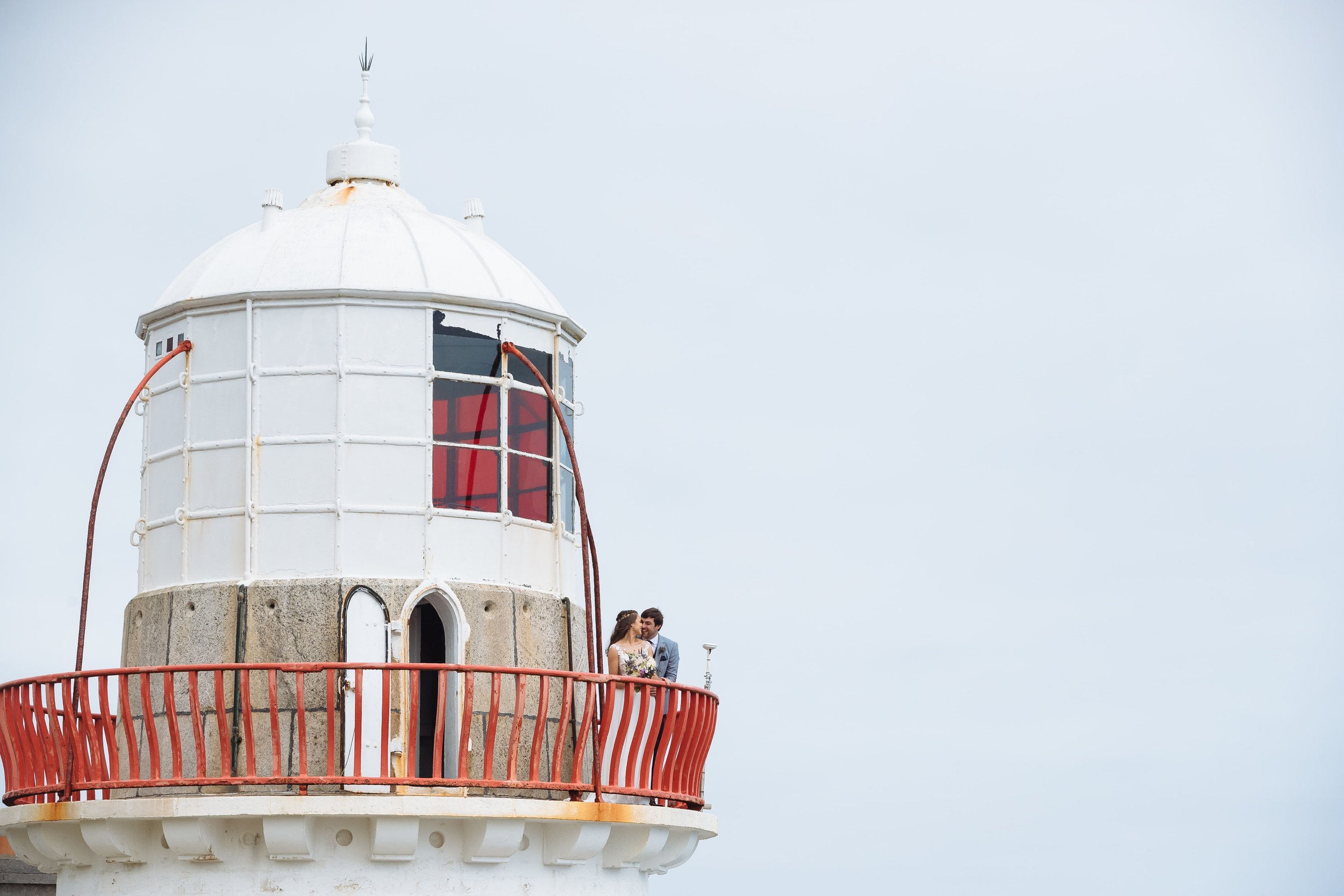 A couple dressed in wedding attire standing on the balcony of a lighthouse with a white tower and a red railing, looking at each other.